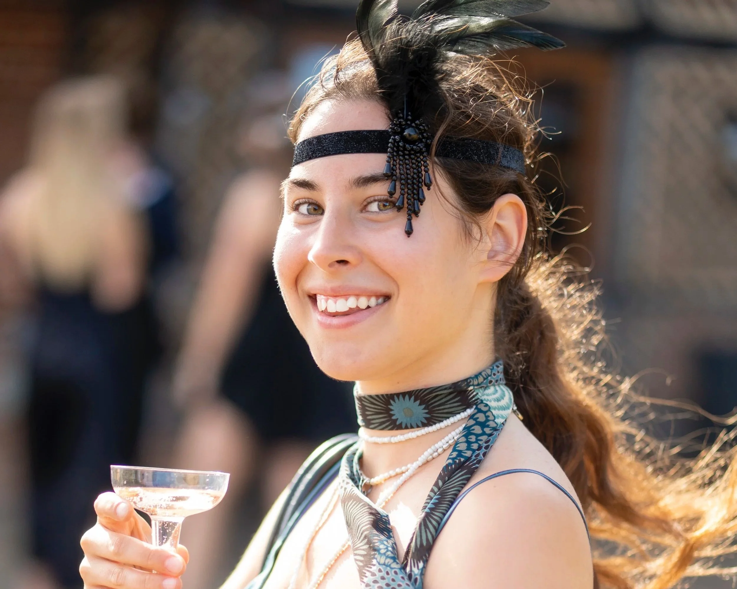 Young woman dressed in vintage 1920s fashion holding a champagne glass, smiling outdoors during daytime.