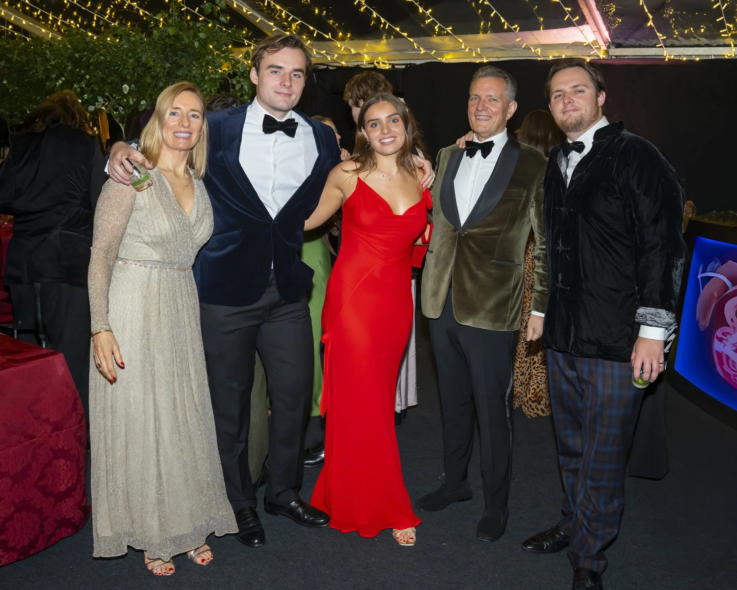 Group of six people dressed in formal attire at an indoor party or event, standing together and smiling. The background features string lights and decorations.