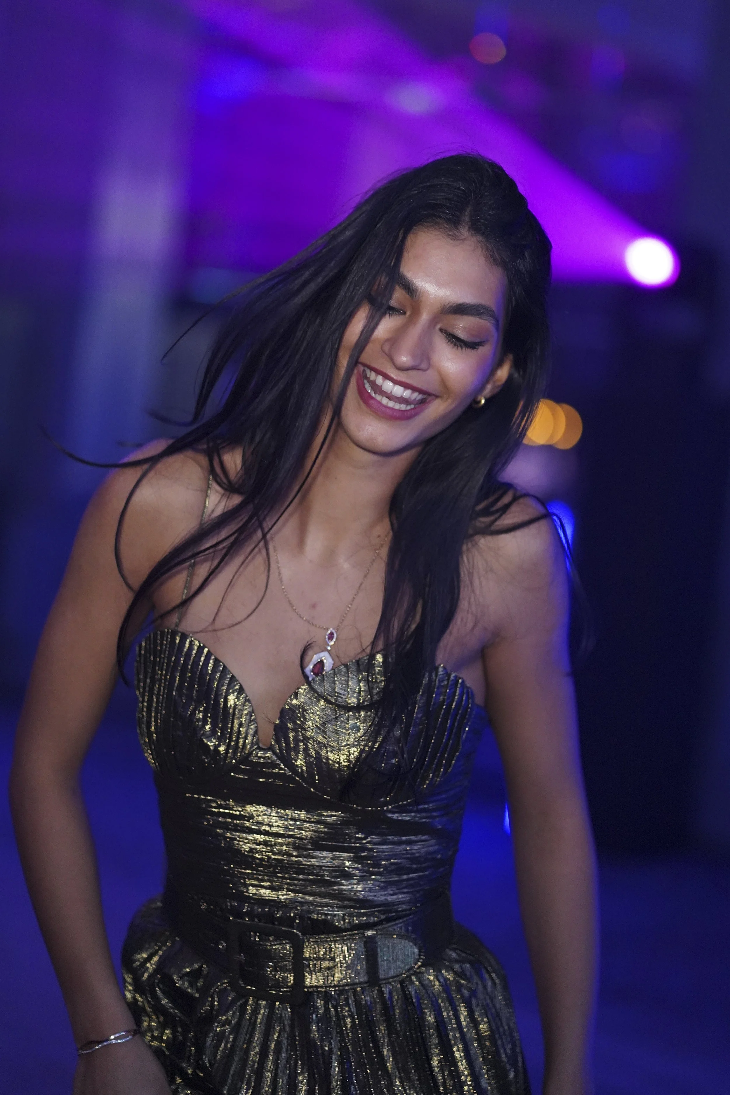 A woman with long dark hair, smiling with eyes closed, wearing a metallic gold and black strapless dress and a necklace, in a setting with purple and blue lighting.
