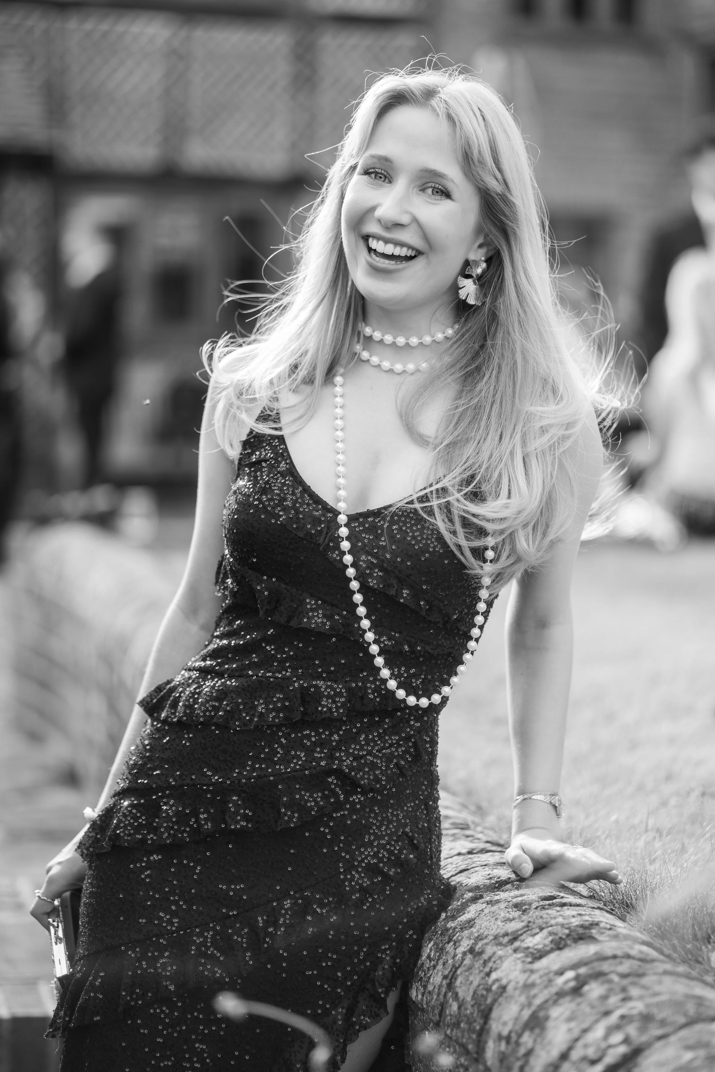A smiling woman with long hair wearing a black, shiny, sleeveless dress, layered pearl necklaces, and earrings, outdoors near a log, with a blurred background.