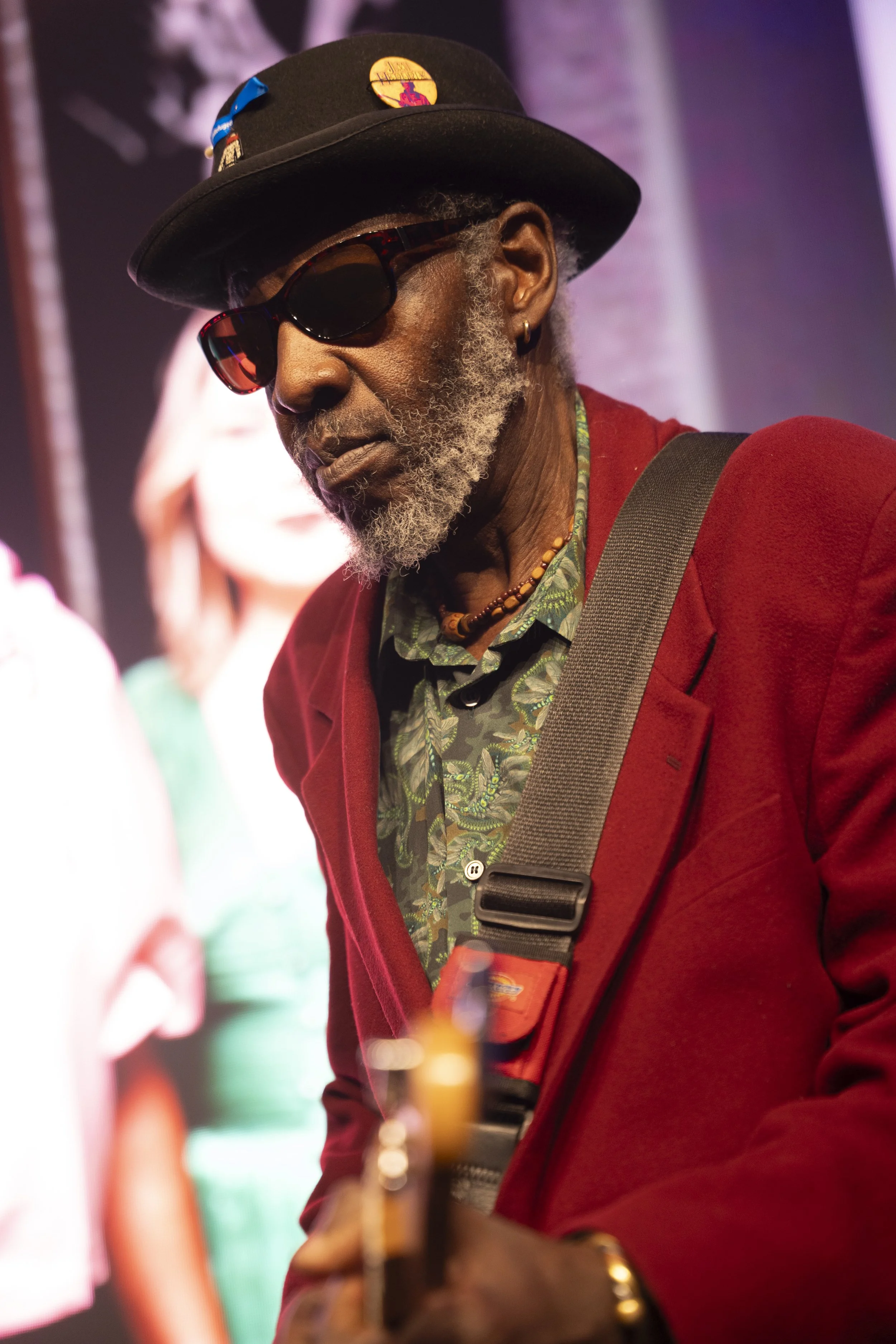 An older man with a gray beard, sunglasses, and a hat decorated with pins, wearing a red jacket over a patterned shirt, appears to be playing guitar.