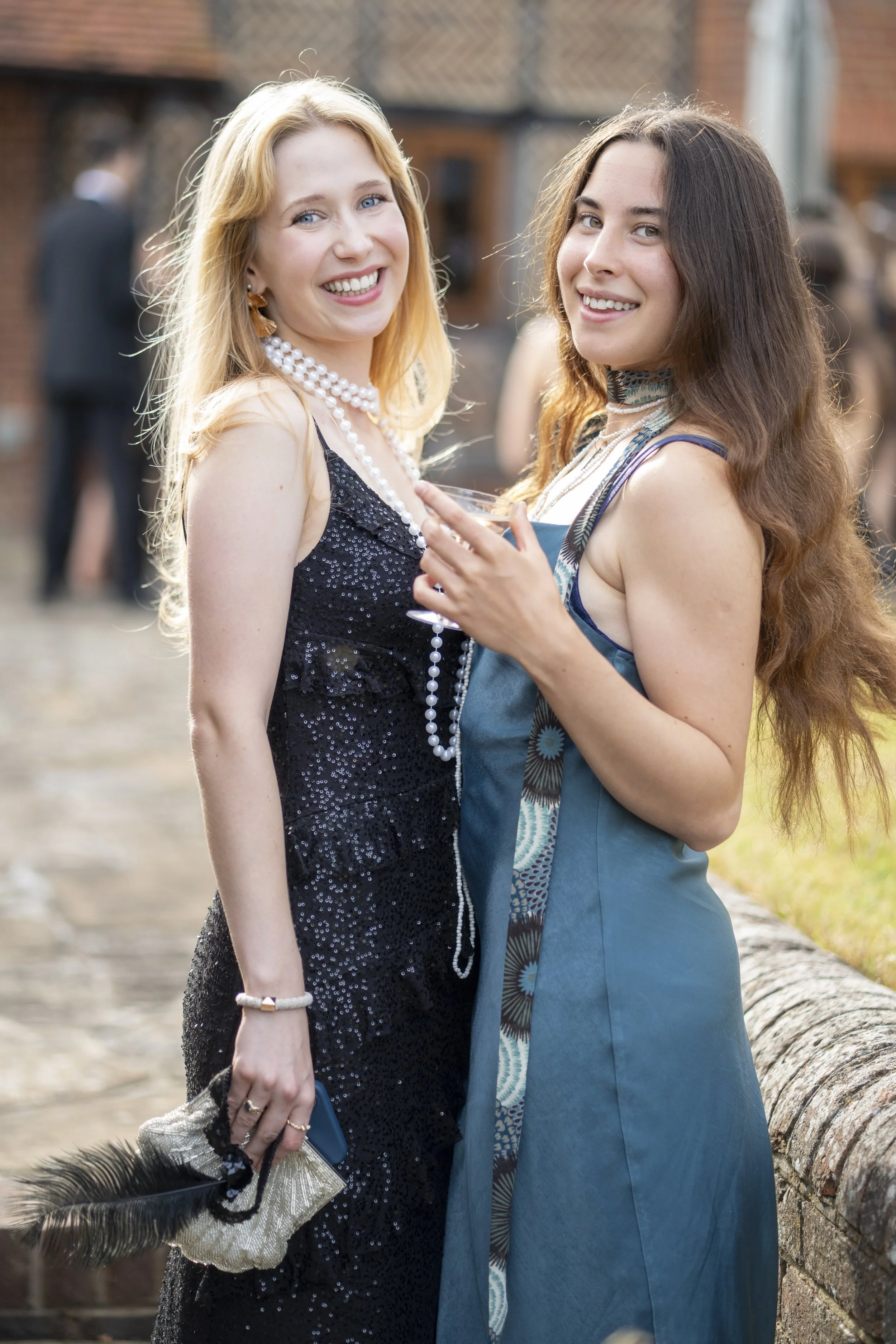 Two women in evening dresses smiling at a social gathering outdoors, with other guests in the background.