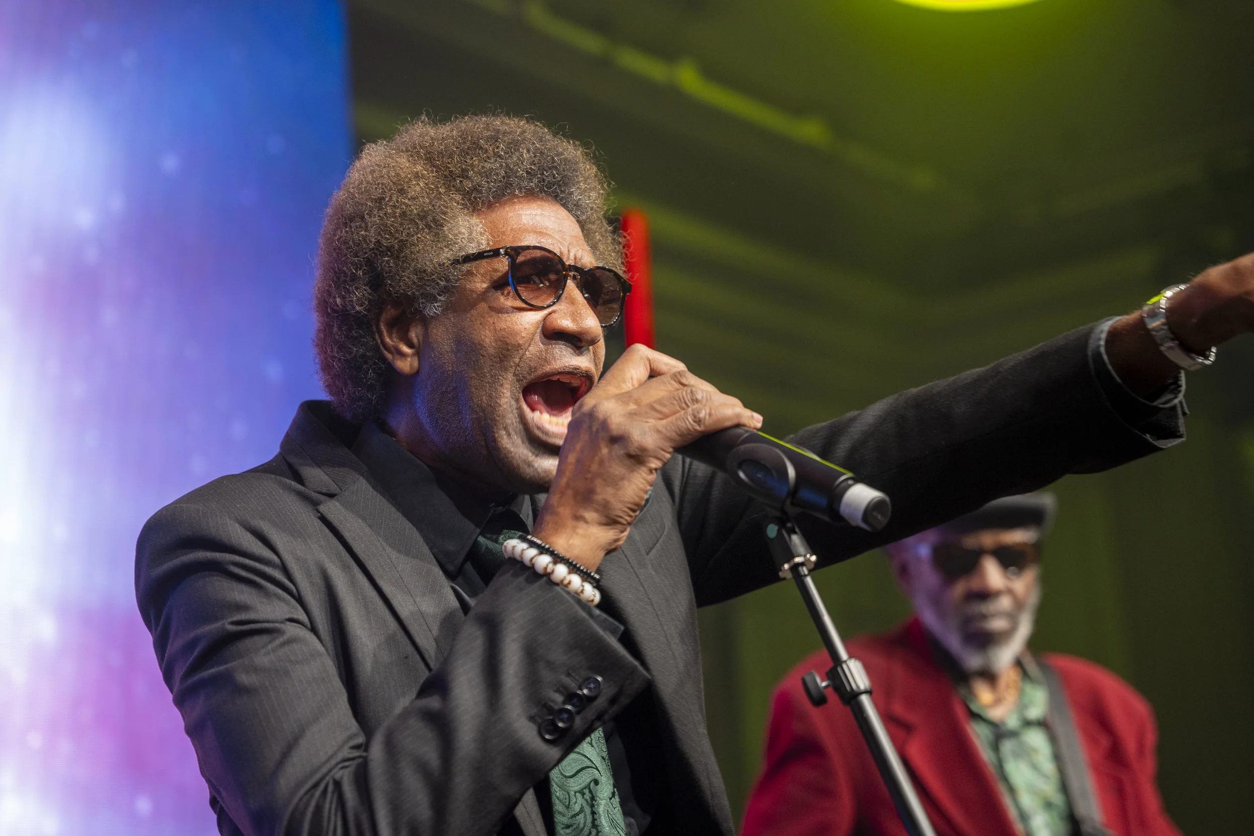 An elderly African American man with glasses, gray hair, and a beard speaking into a microphone on stage. He's wearing a dark suit with a green tie and is gesturing with his right hand. Another man with glasses and a gray beard, dressed in a red jack