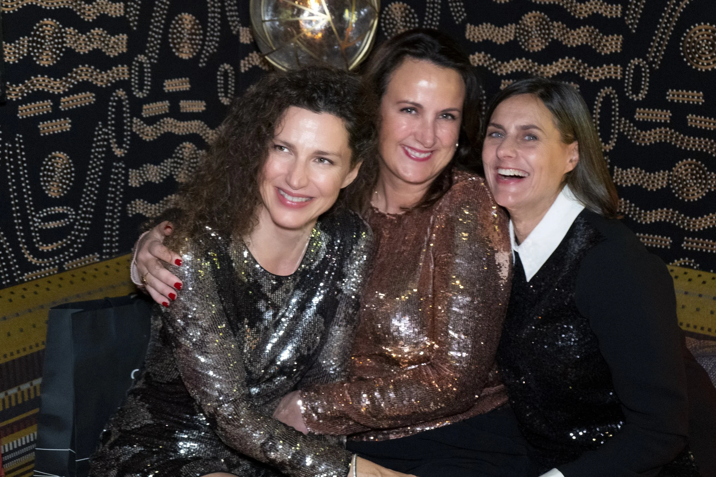Three women smiling and sitting close together at a party or celebration, wearing shiny, sequined tops, with a decorative black and gold background.