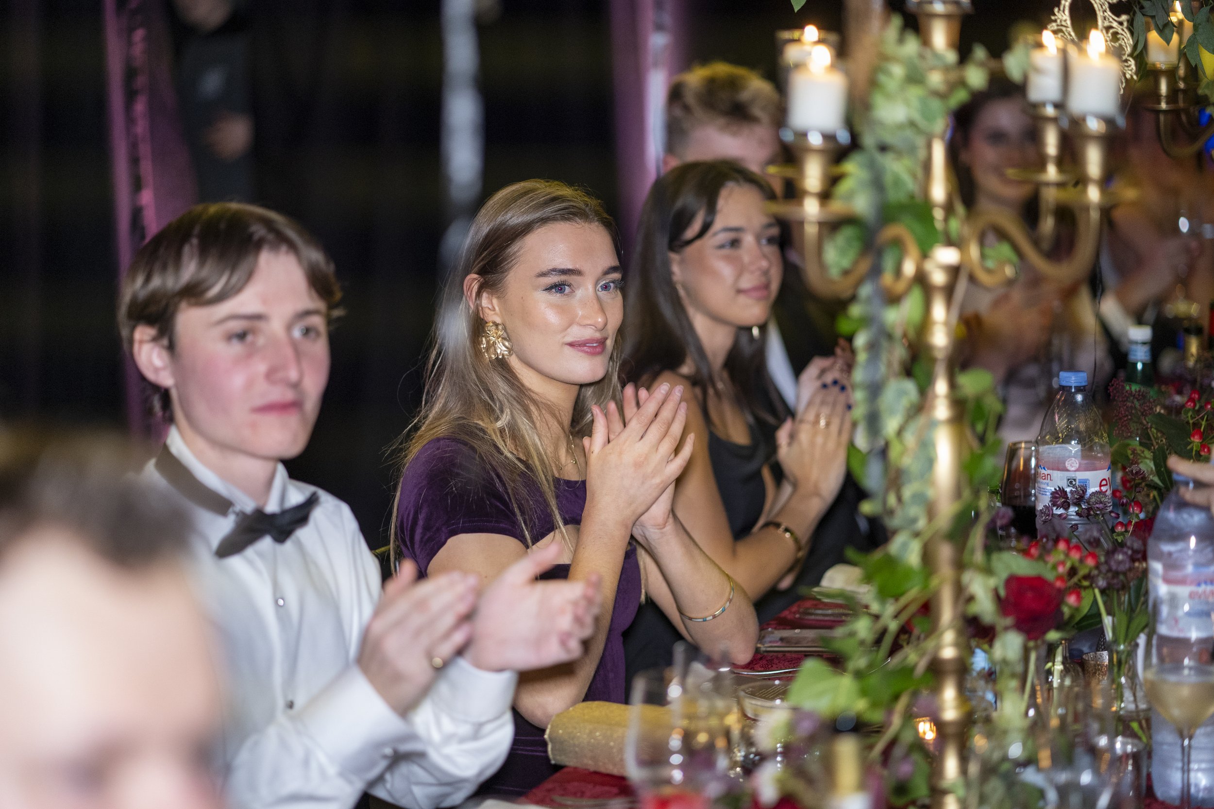 People dressed formally, sitting at a decorated table, clapping, during a celebration or dinner event.