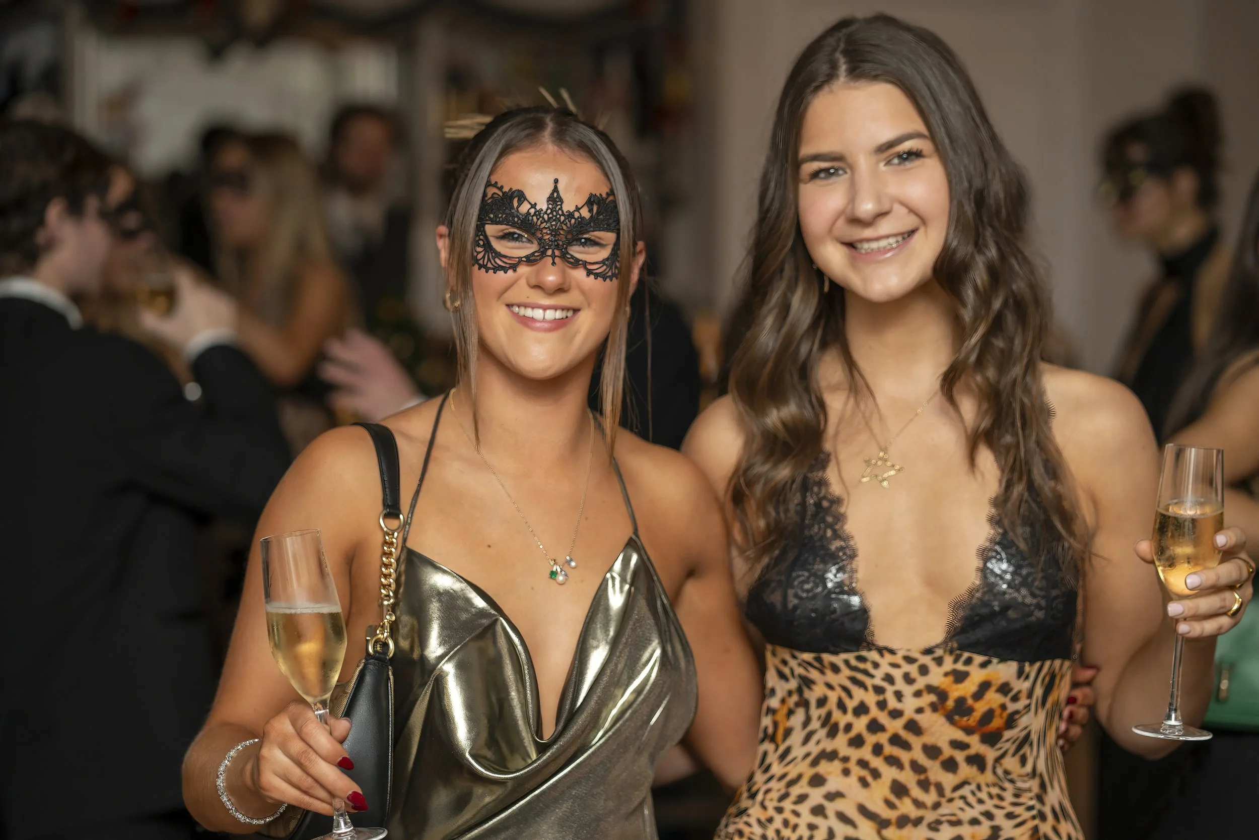 Two women smiling at a party, holding glasses of champagne, dressed in festive outfits, one wearing a black lace mask, in a lively social setting.