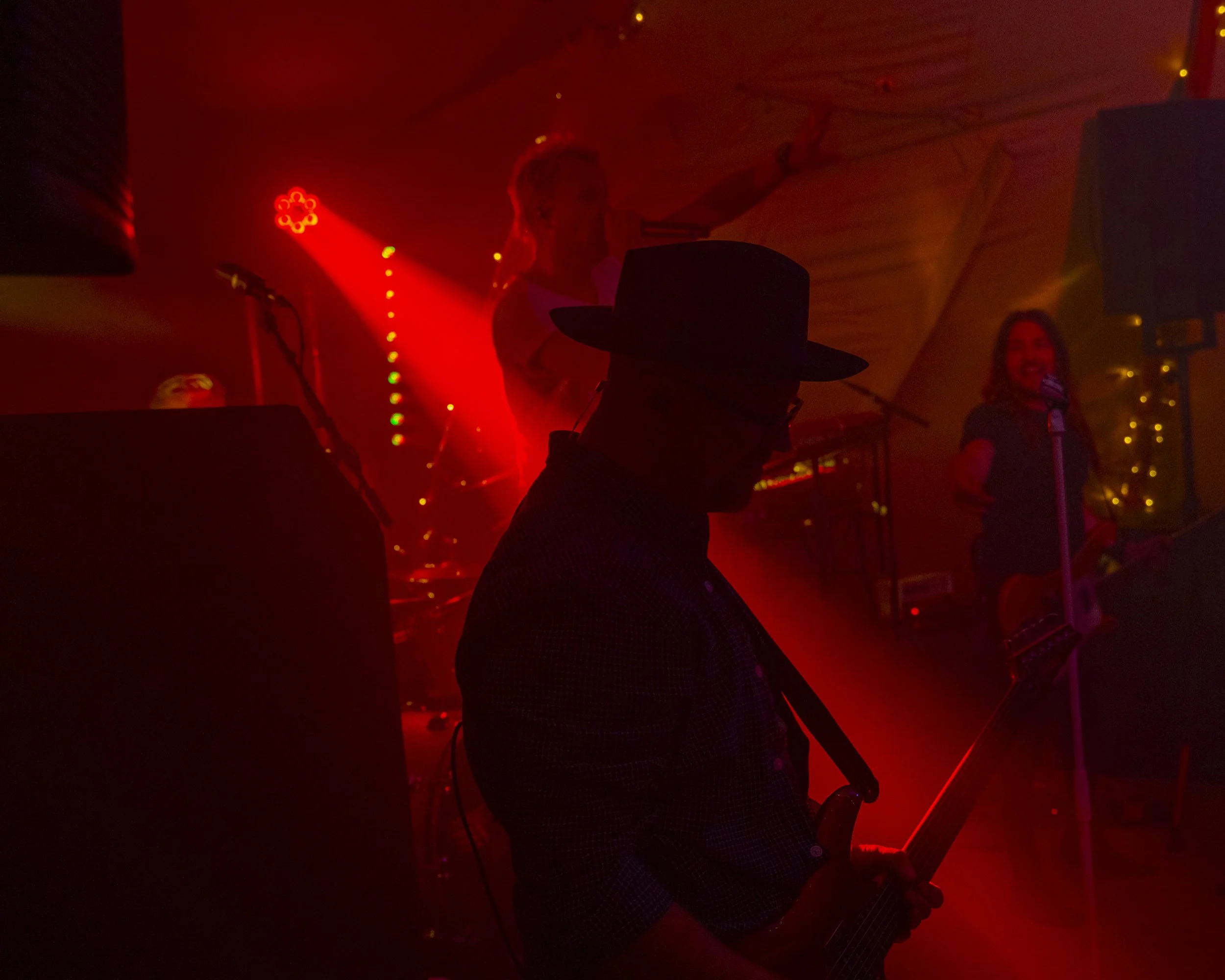 Silhouetted man wearing a hat playing guitar with a band performing in a red-lit scene with a woman singing into a microphone.