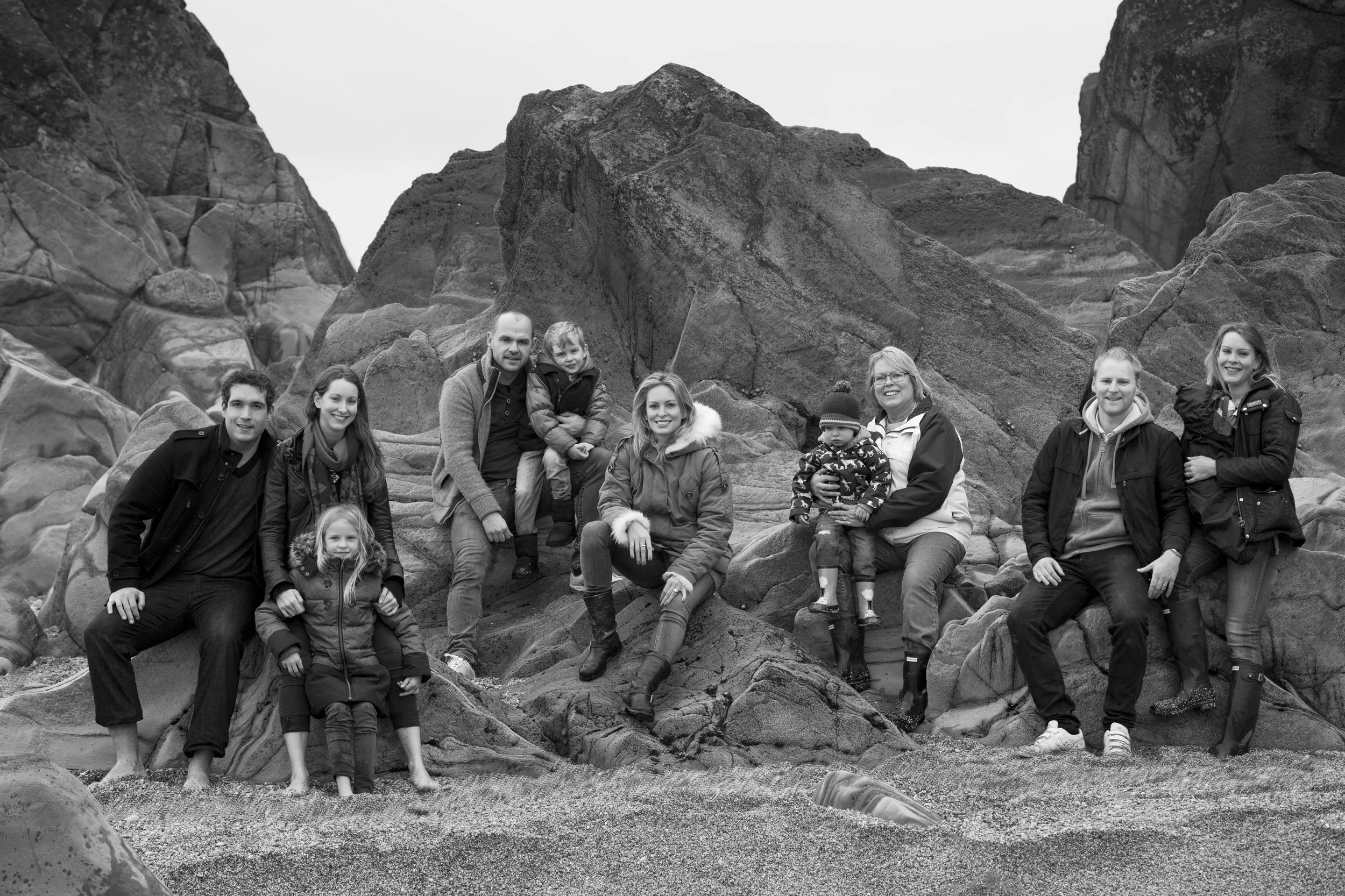 A black and white photo of a family posing among rocks and boulders on a beach. The family includes adults and children, all dressed warmly in jackets and boots, with some sitting on rocks and others standing or crouching. The background shows large 