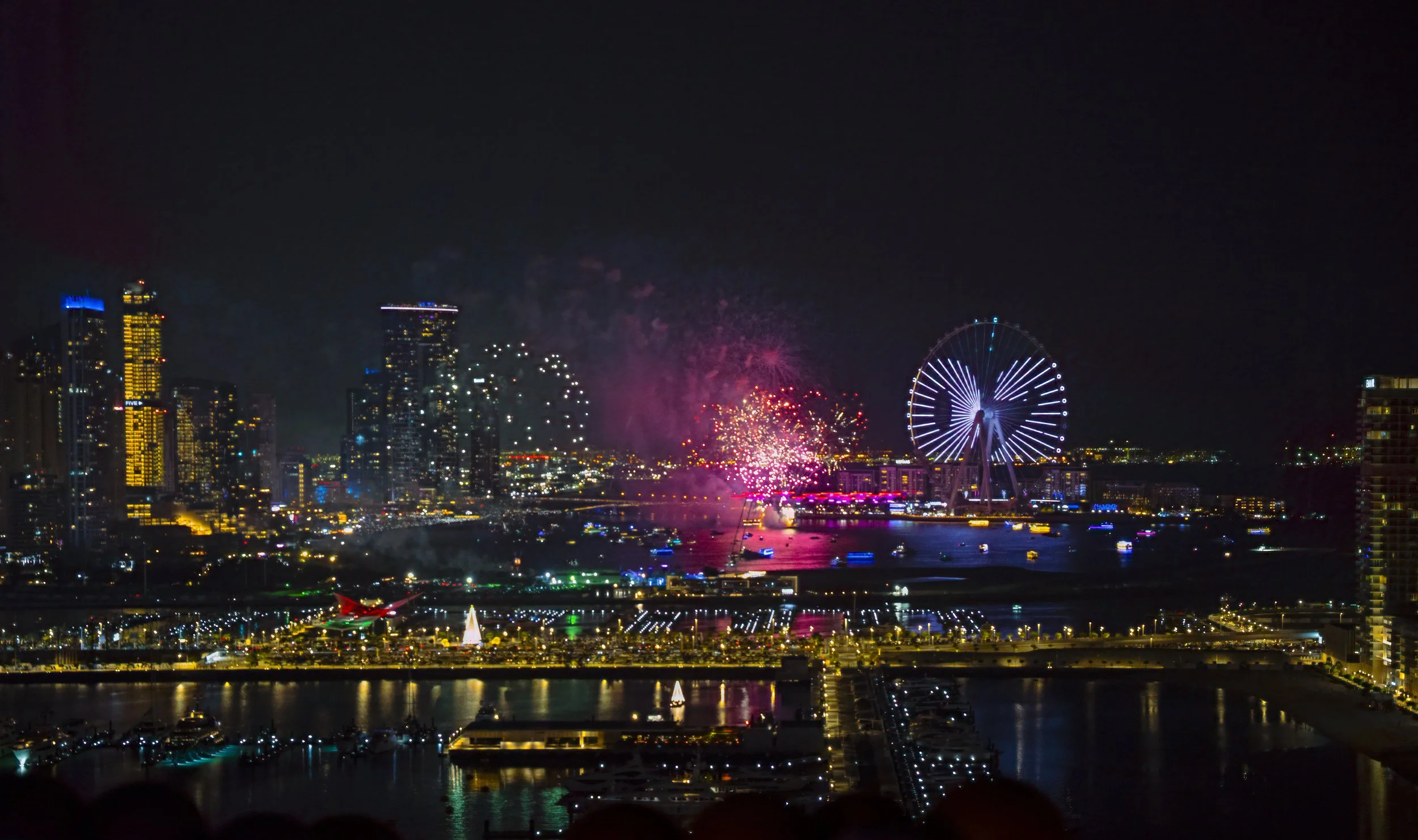 Dubai Nighttime cityscape with fireworks, ferris wheel, and illuminated skyline reflected in water.
