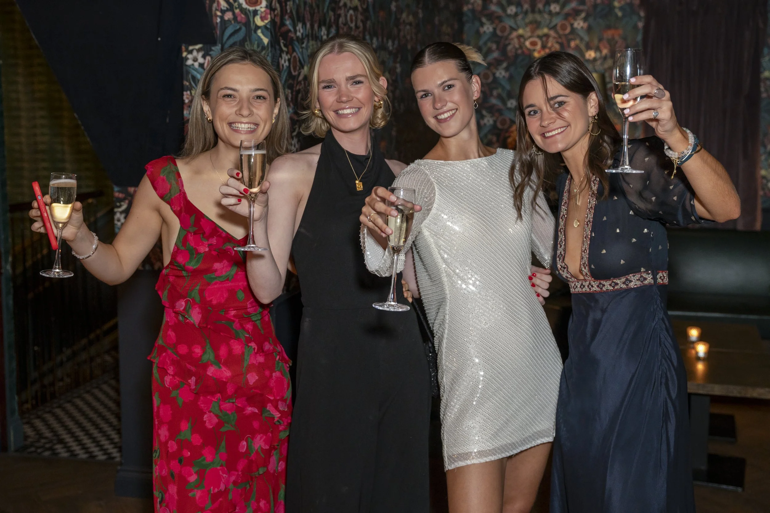 Five women dressed in formal attire celebrating with glasses of champagne at an indoor party.