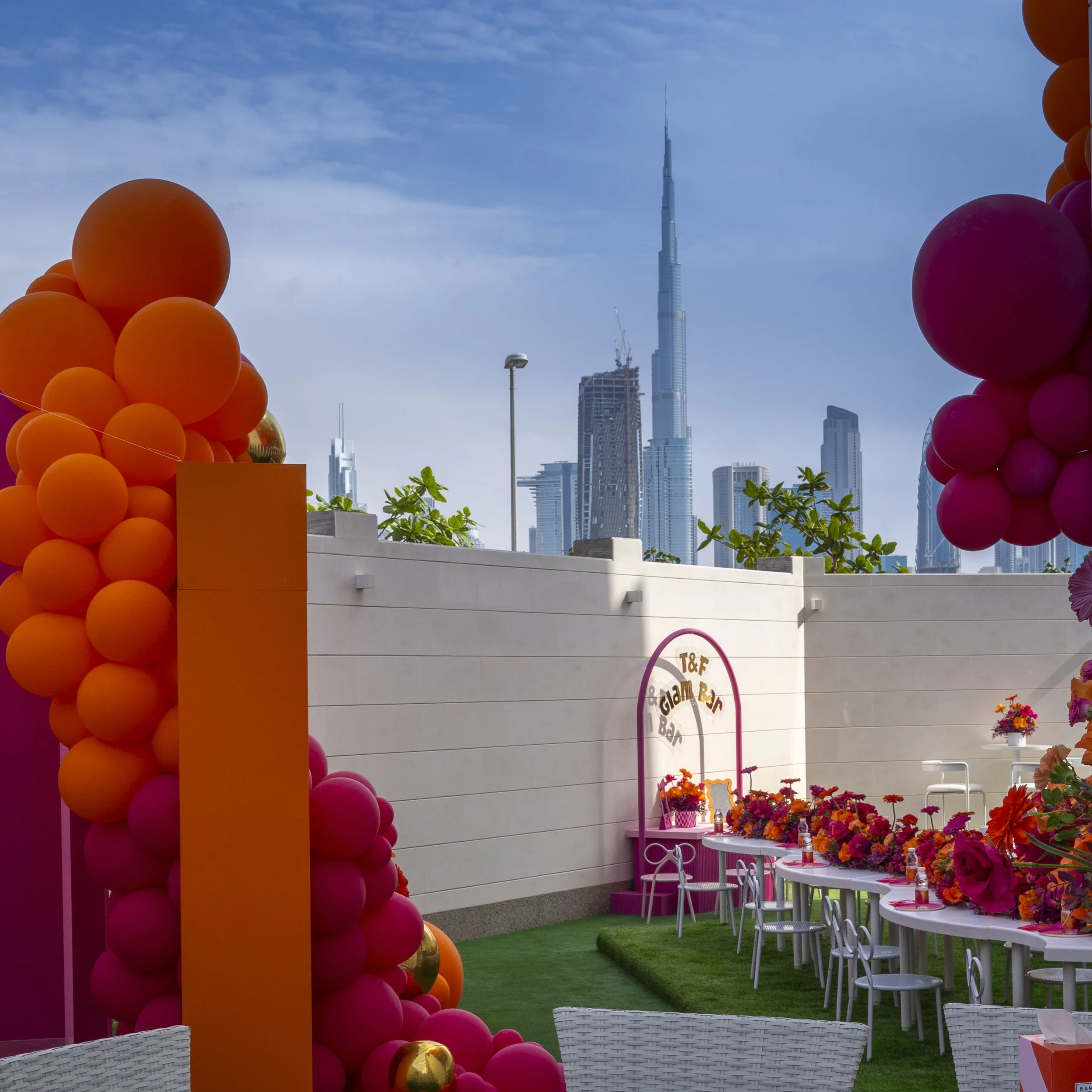 Dubai Party Decorated outdoor space with pink and orange balloons, white tables with floral arrangements, and a city skyline including the Burj Khalifa in the background.