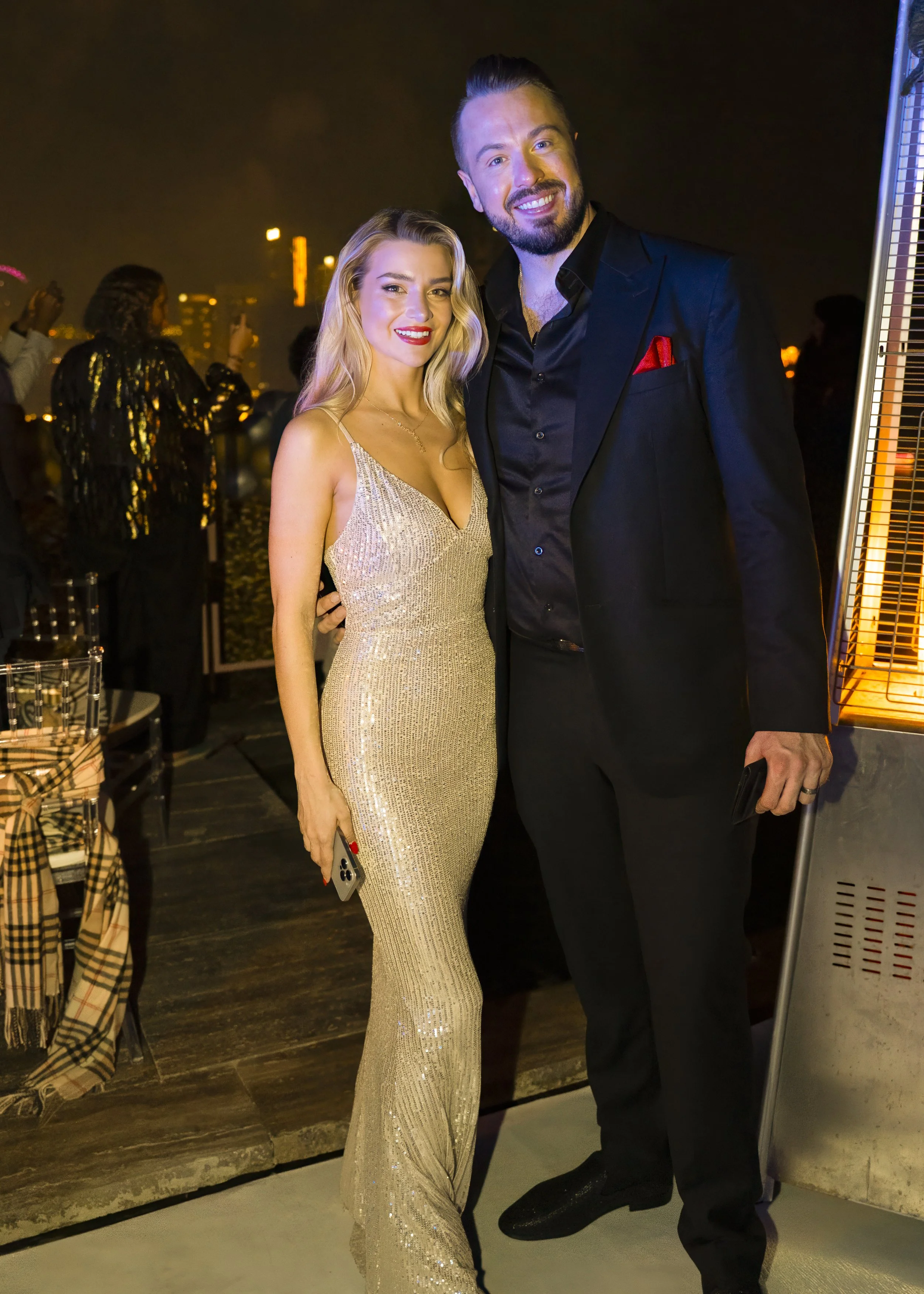 A famous magician and his assistant woman at a Dubai event. She wears a sparkling gold gown and a man in a black suit pose together at a social event during nighttime, with city lights in the background.