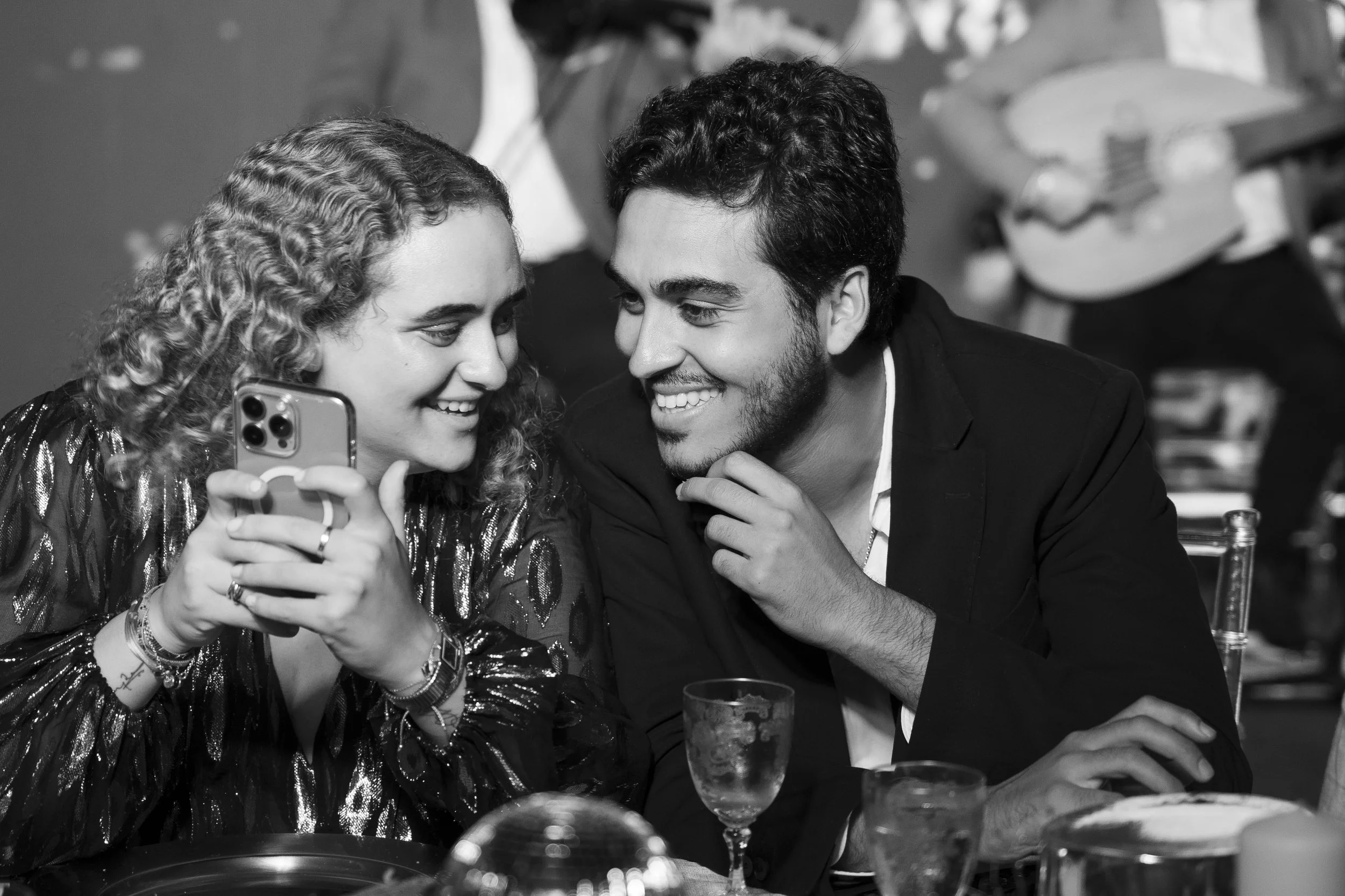 A smiling woman and man sharing a moment at a restaurant, with the woman showing something on her phone, while the man touches his chin, surrounded by drinks and dessert.