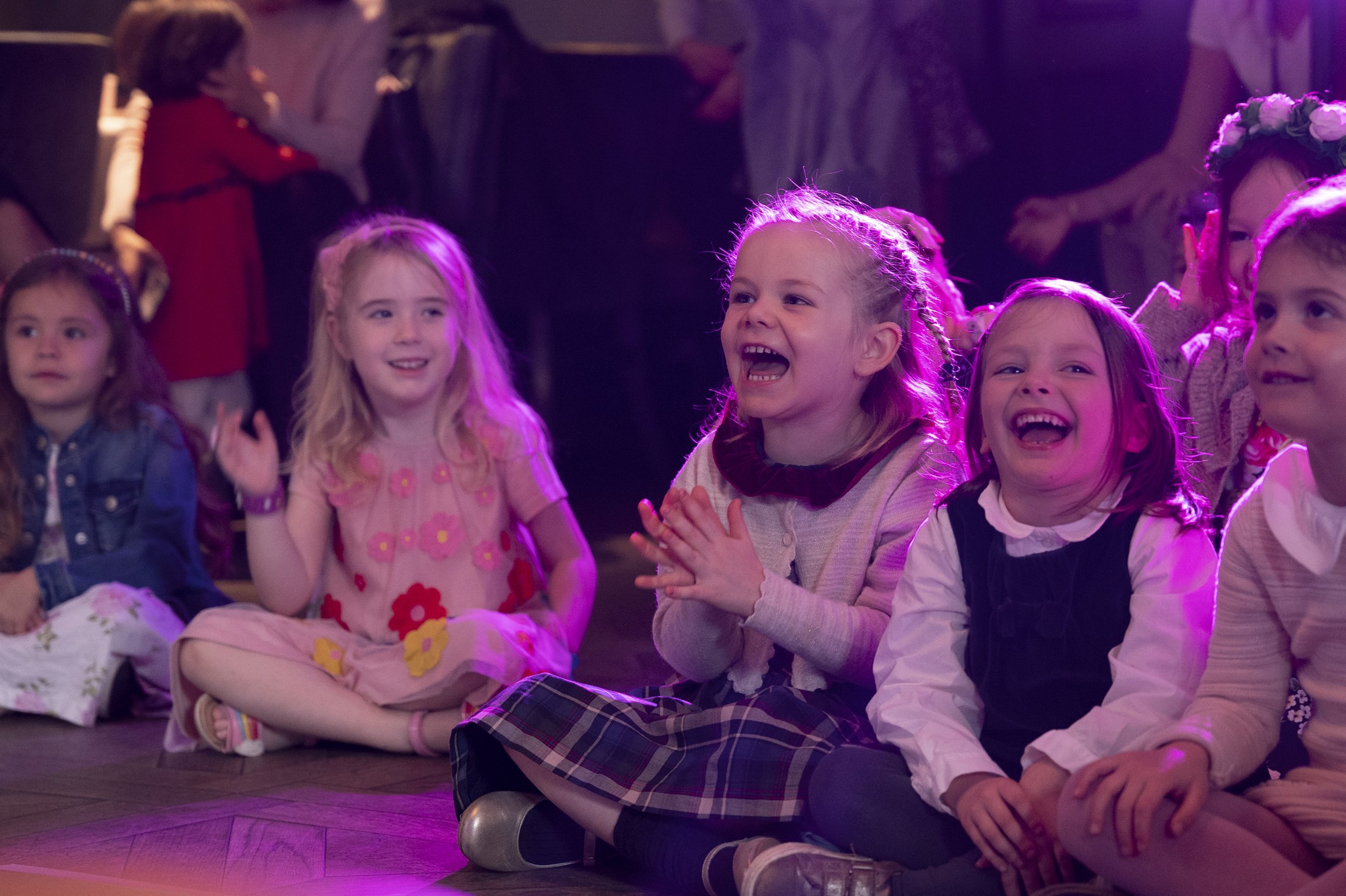 Group of young children sitting on the floor, laughing and enjoying a performance or event.