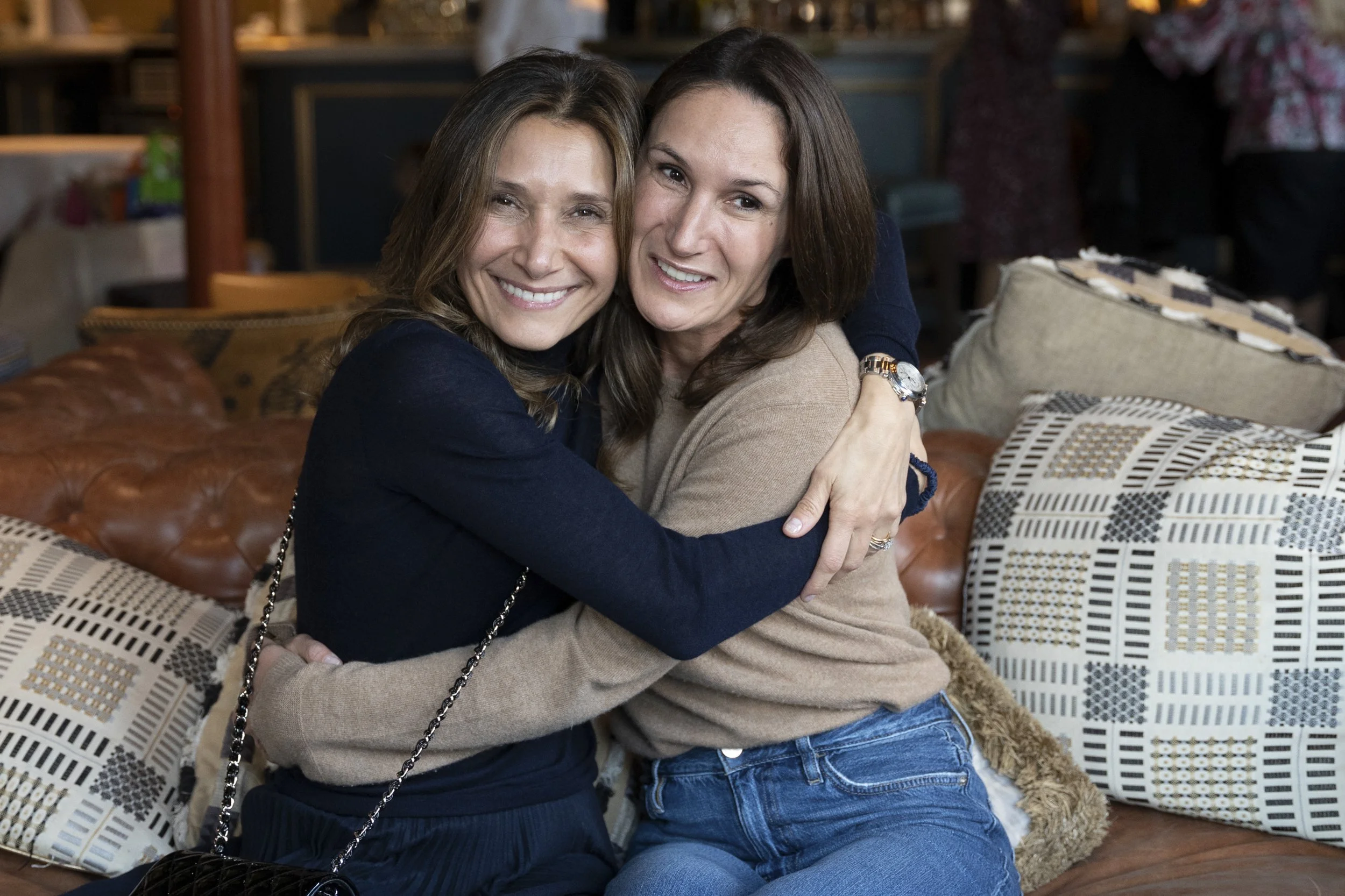 Two women hugging and smiling on a couch in a cozy indoor setting, with decorative pillows around them.