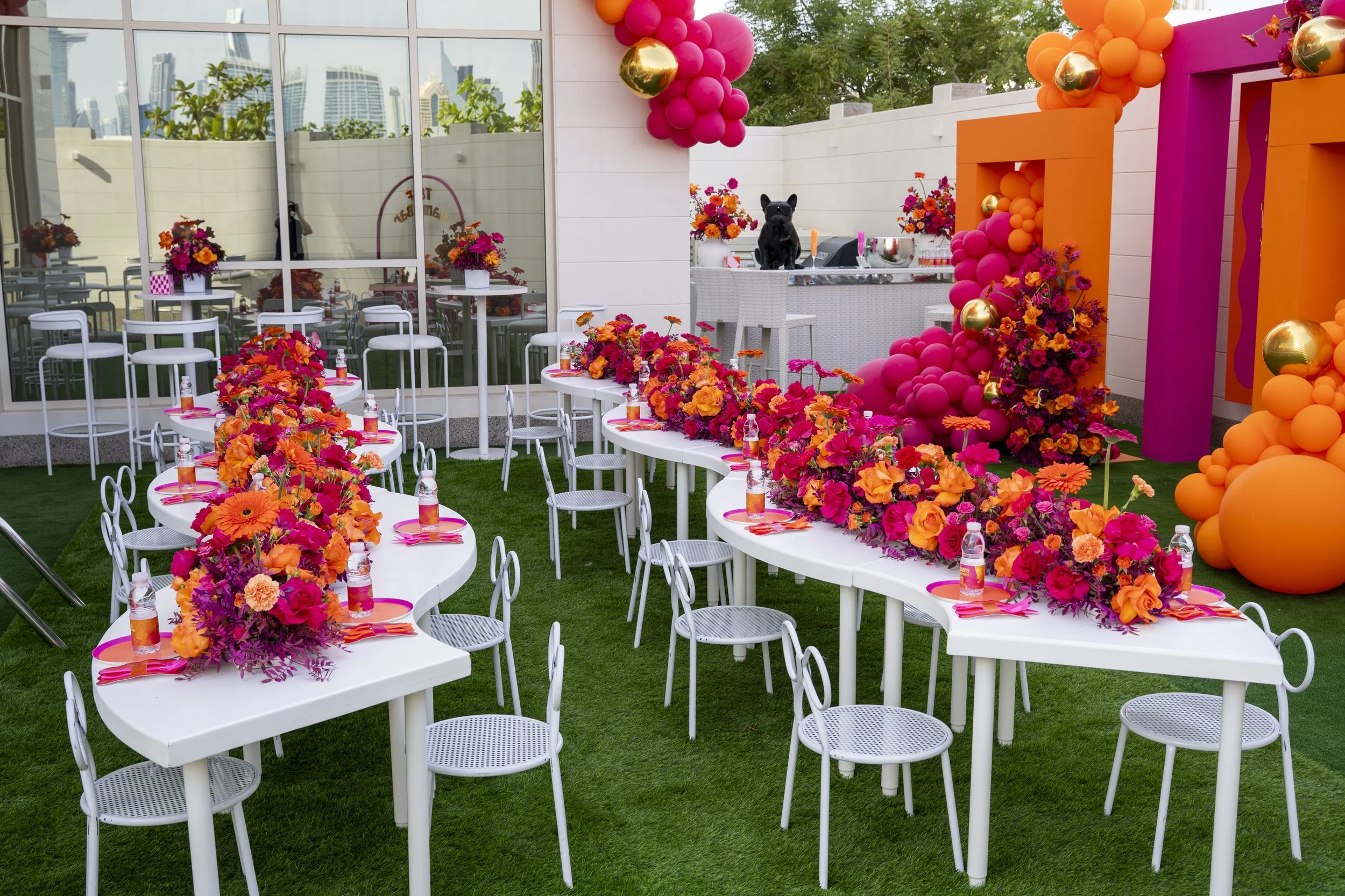Decorated outdoor party area with white tables, colorful pink and orange floral centerpieces, pink and orange balloons, and a bar with black French bulldog statues, set on artificial green grass.