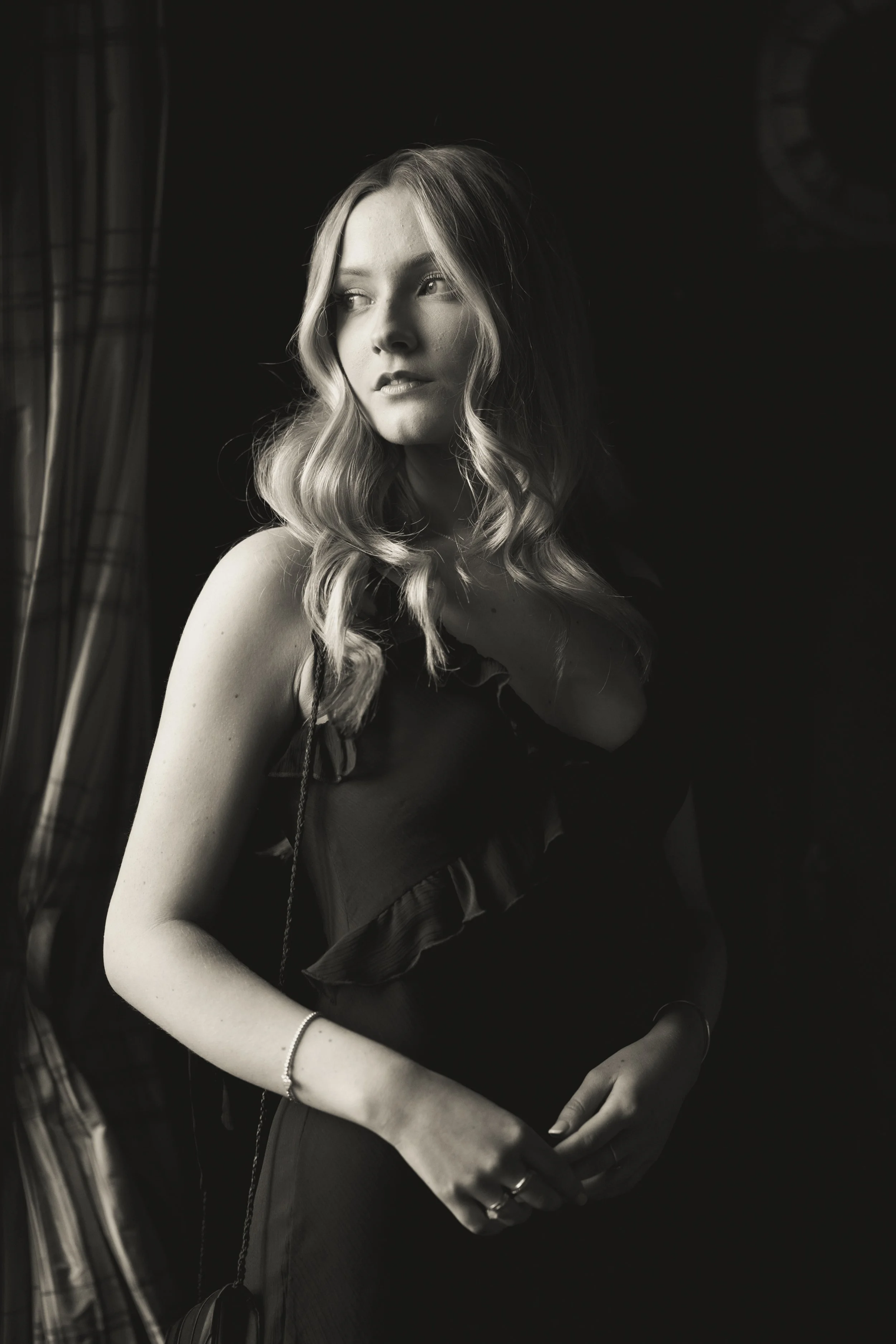 A black-and-white photo of a young woman with wavy hair, wearing a dress with ruffled straps, standing near a window with curtains, looking away from the camera with a thoughtful expression.
