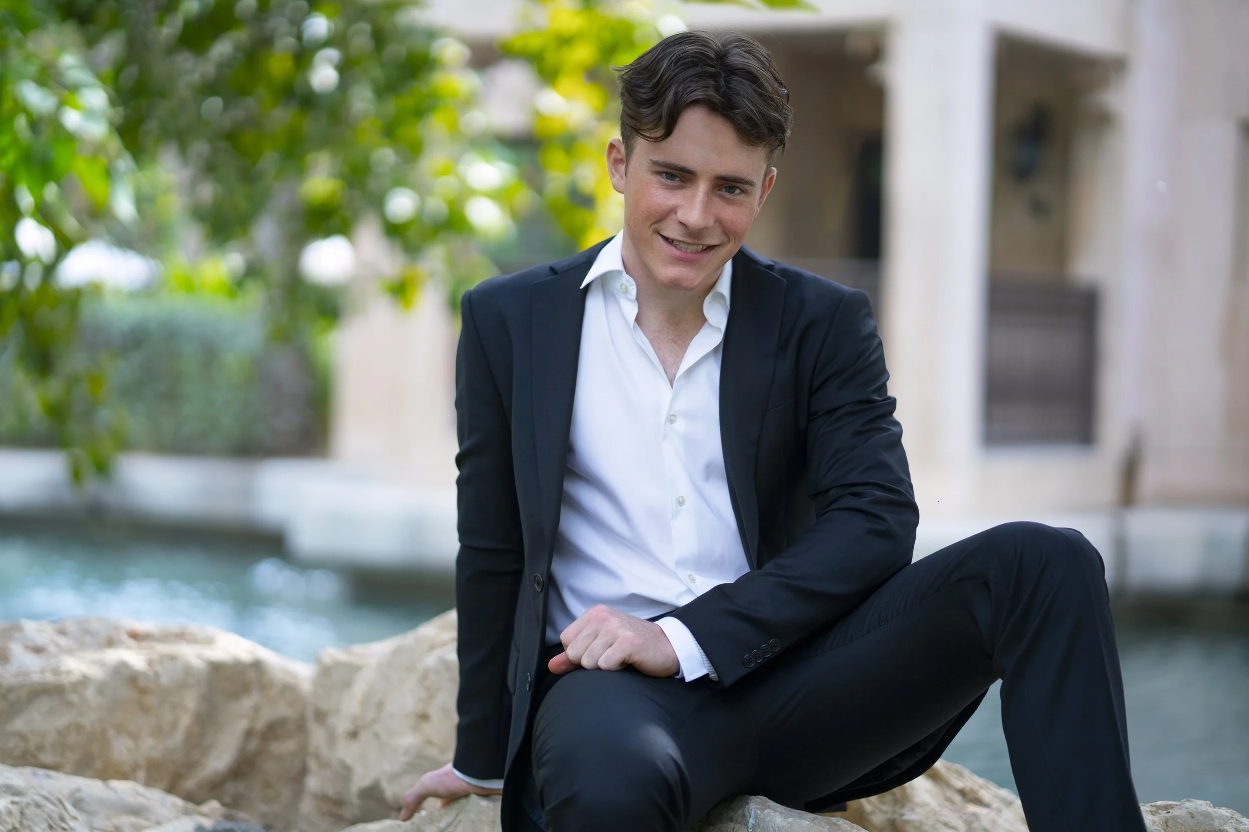 A young man in a black suit and white shirt sitting on rocks outdoors near water, smiling at the camera.