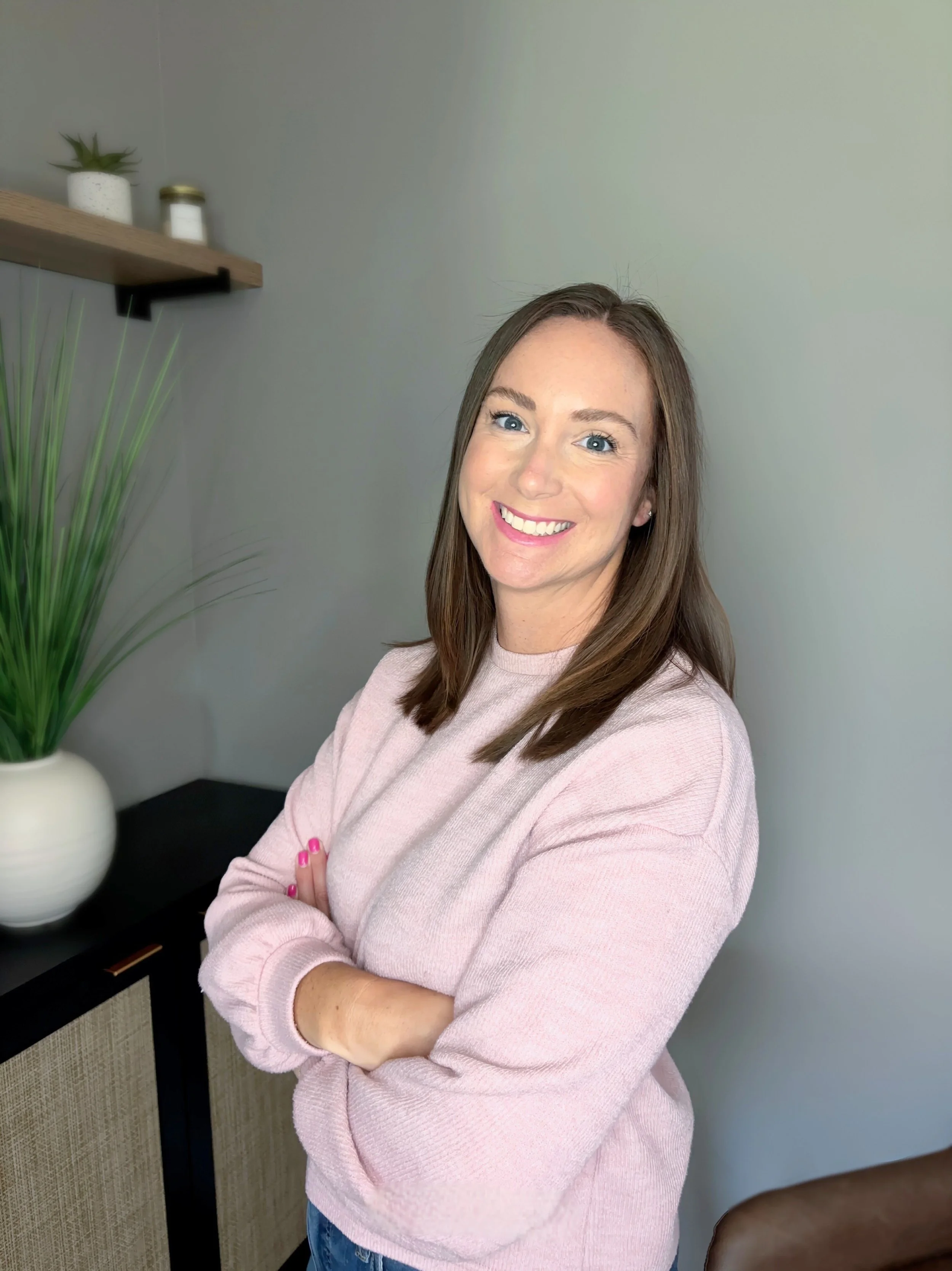 A smiling woman with shoulder-length brown hair, wearing a pink sweater, stands indoors with her arms crossed. Behind her is a gray wall, a wooden shelf with small potted plants and jars, and a black cabinet with beige panels.