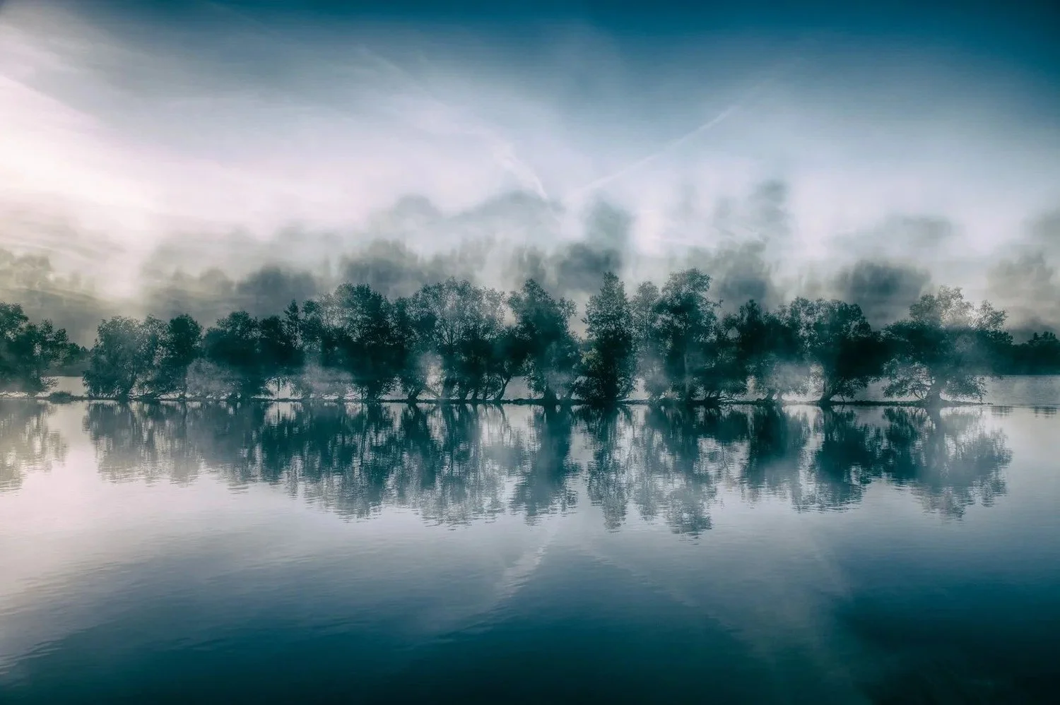 Morgendliche Szene mit Nebel über einem ruhigen Fluss und Bäumen, die sich im Wasser spiegeln.