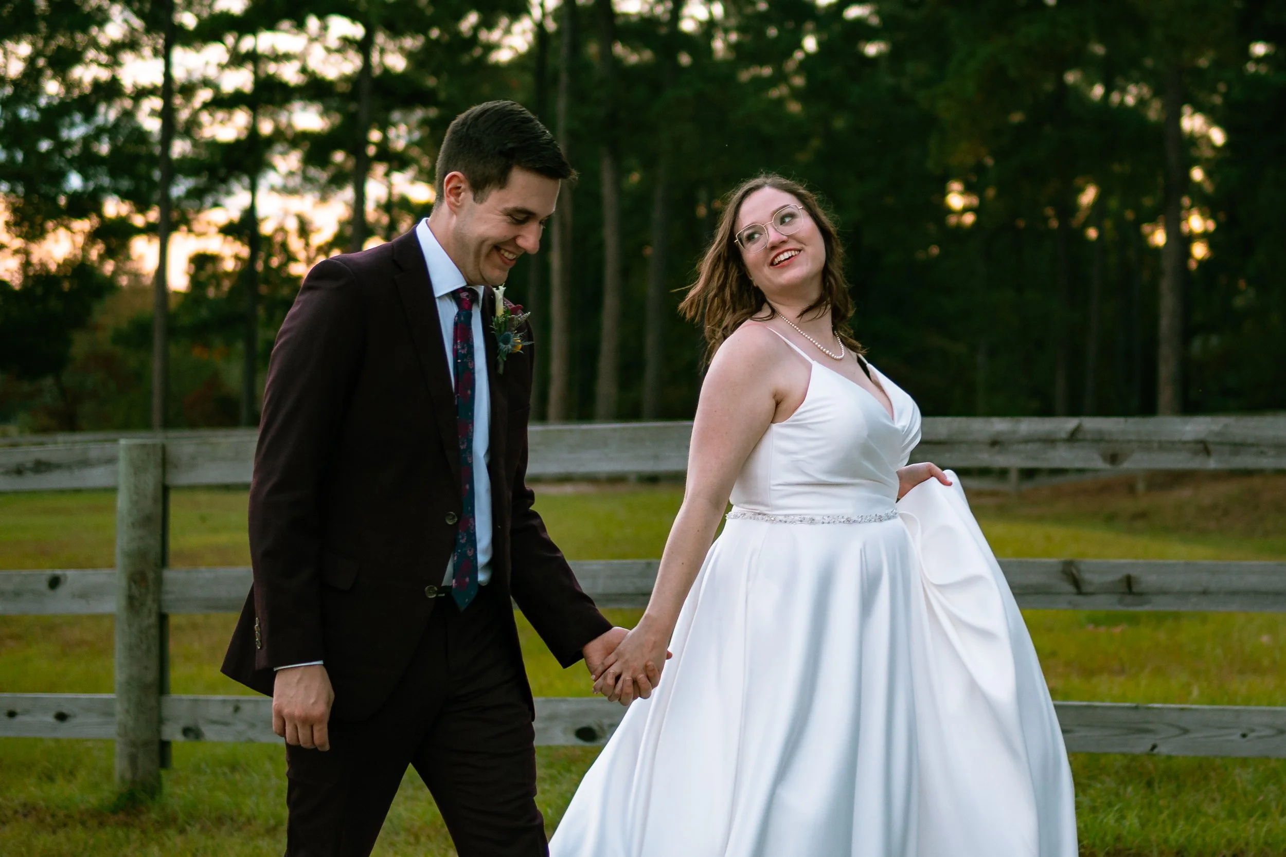 A couple walking hand in hand outdoors at sunset; the man is dressed in a suit with a boutonniere, and the woman is in a white dress with pearls, smiling at each other.
