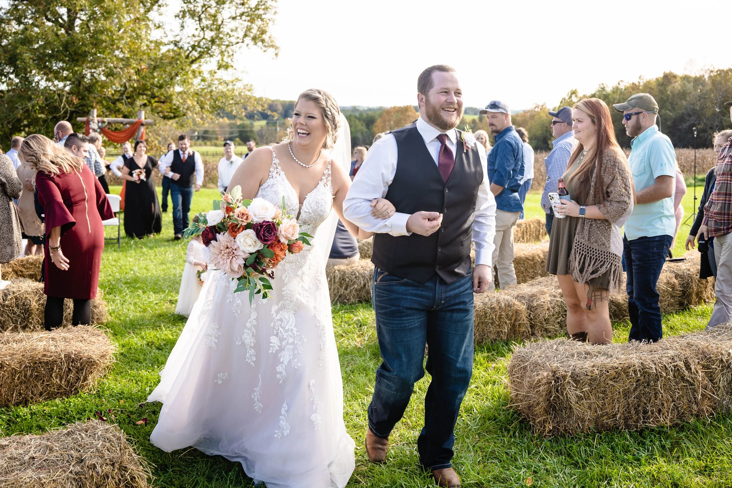A bride and groom smile as they walk outdoors on their wedding day, surrounded by guests and hay bales on a farm with trees in the background.