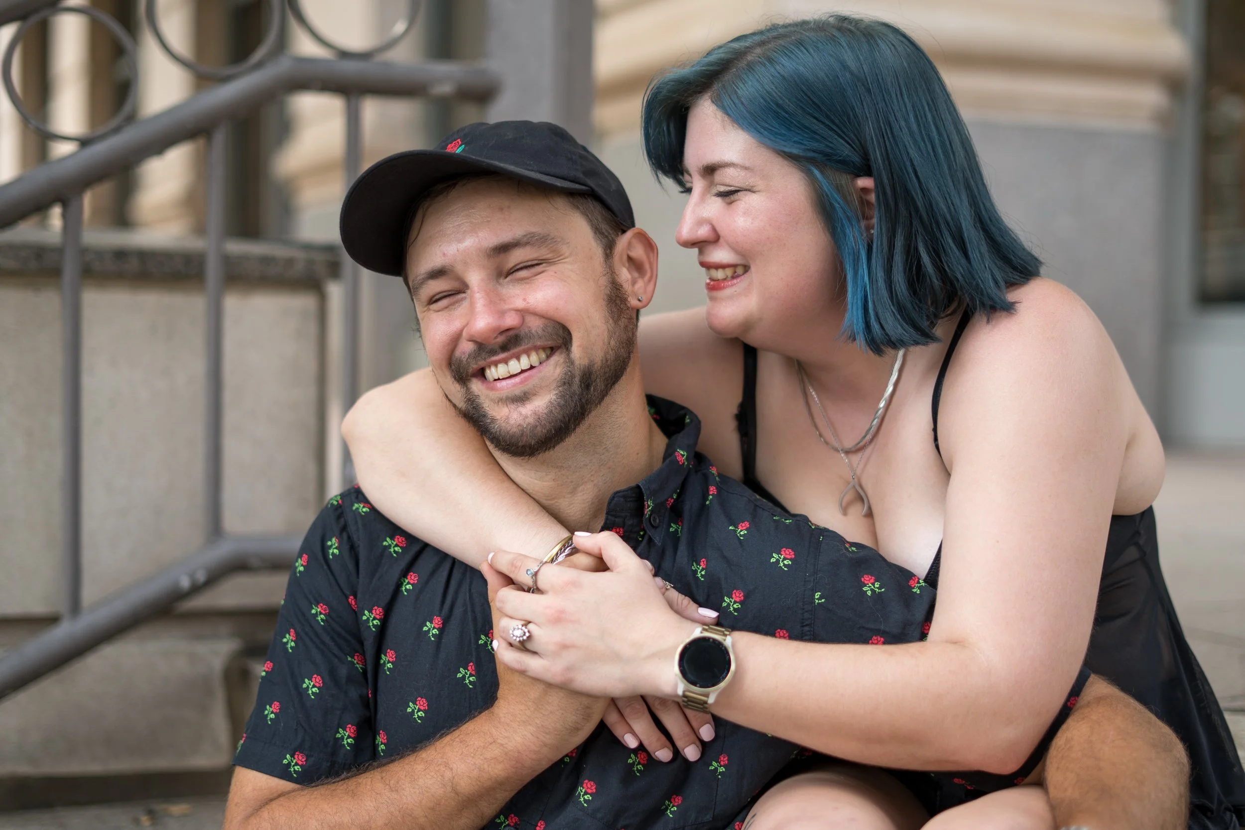 A smiling man and woman hugging each other outdoors on a staircase. The woman has blue hair and the man is wearing a black cap and a black shirt with a floral pattern.