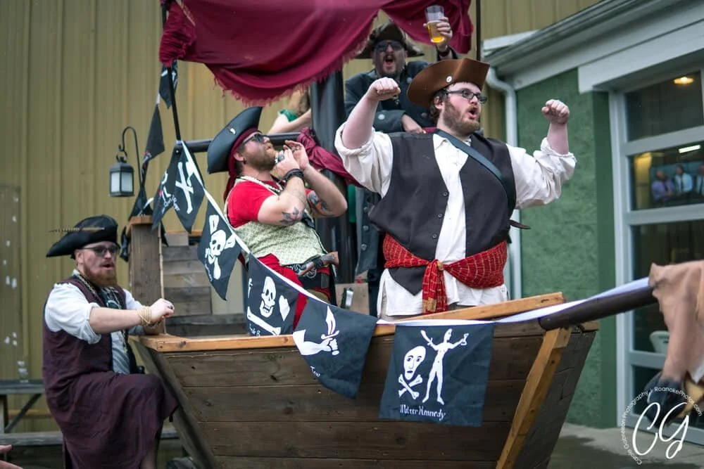 People dressed as pirates on a decorated wooden boat, with some raising their fists and holding drinks, celebrating outdoors.