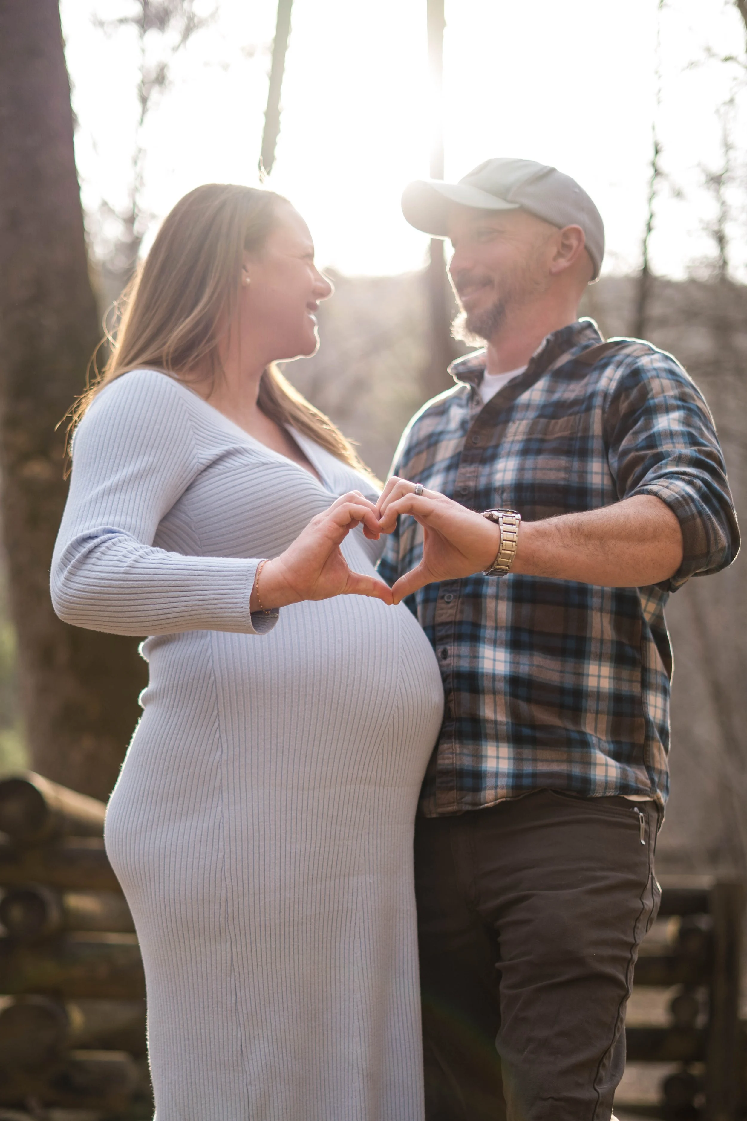 Expectant parents are creating a heart with their hands during golden hour in the woods