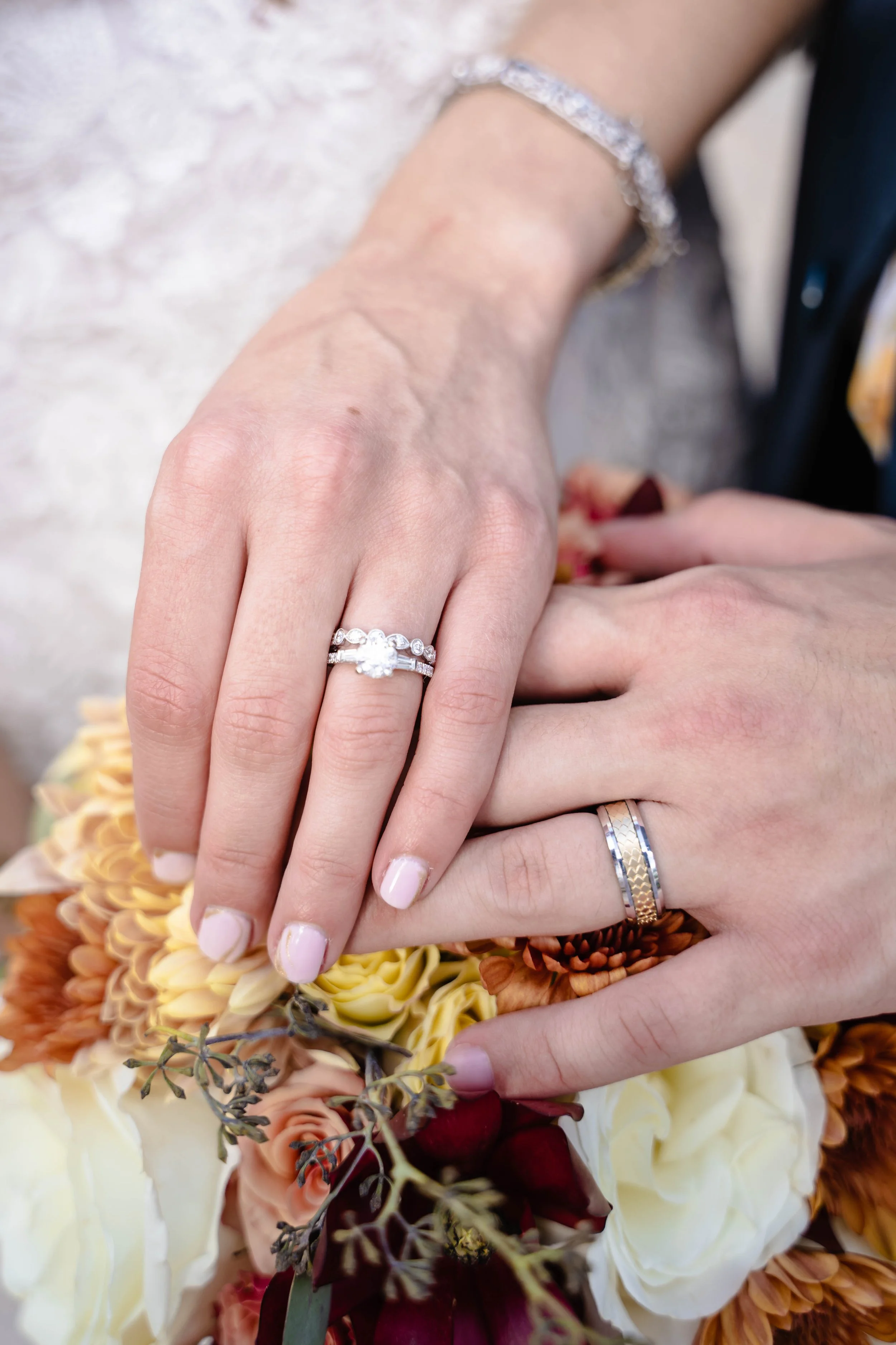 Two hands with wedding rings resting on a bouquet of flowers. The woman's hand has a diamond engagement ring and an additional band. The background shows various flowers in shades of white, orange, burgundy, and yellow.