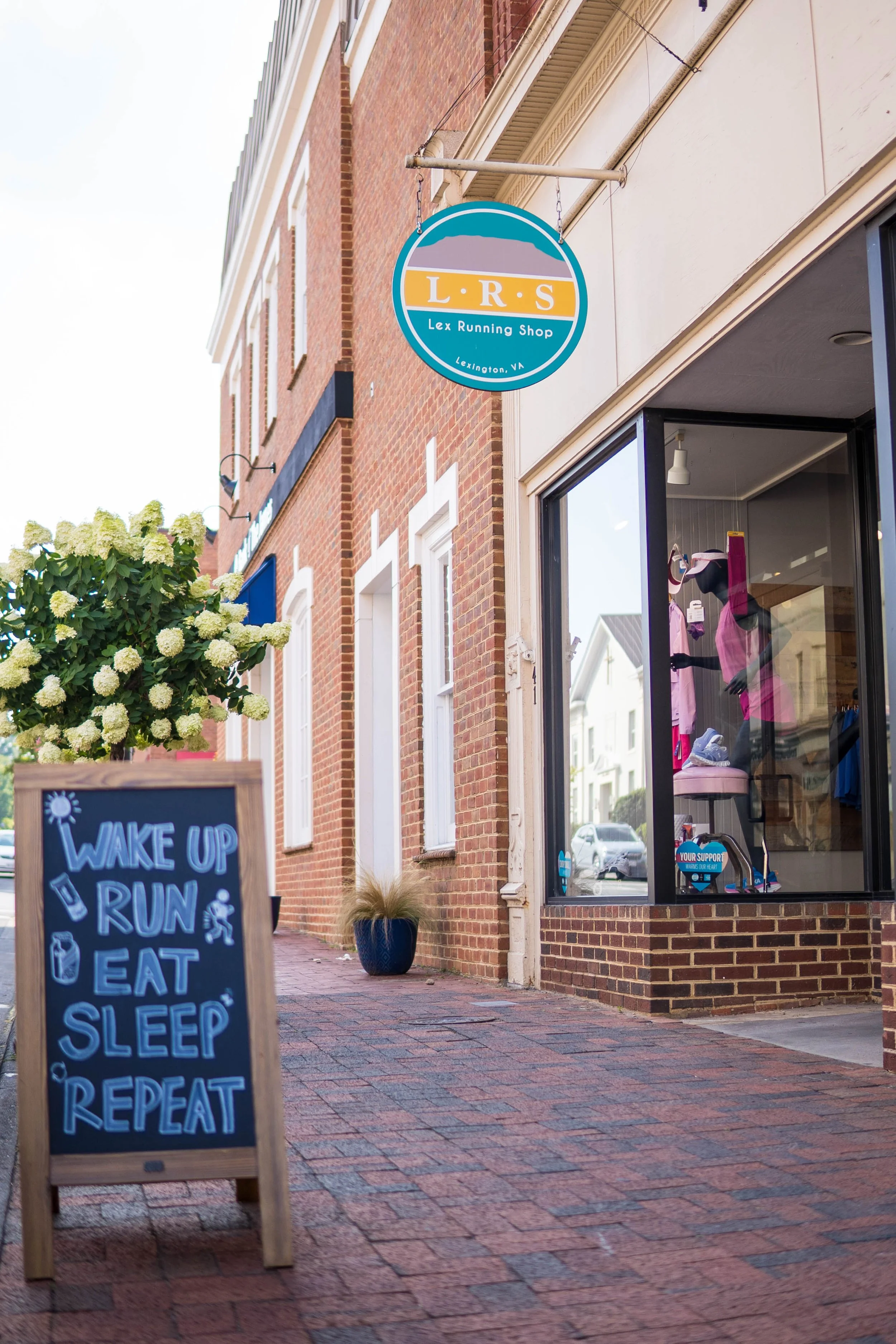 Storefront with a sign that reads 'LRS Lex Running Shop Lexington VA' and a chalkboard sign outside that says 'Wake Up Run Eat Sleep Repeat' on a brick sidewalk.
