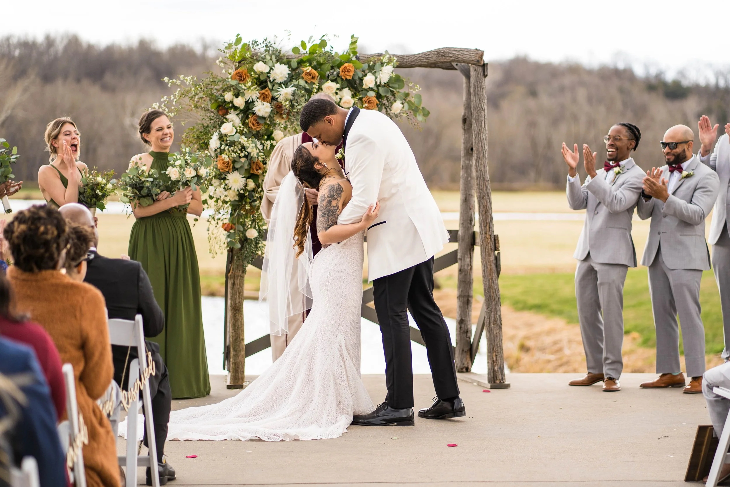 Couple kissing during outdoor wedding ceremony with friends and family watching, decorated with floral arch and natural landscape in the background.
