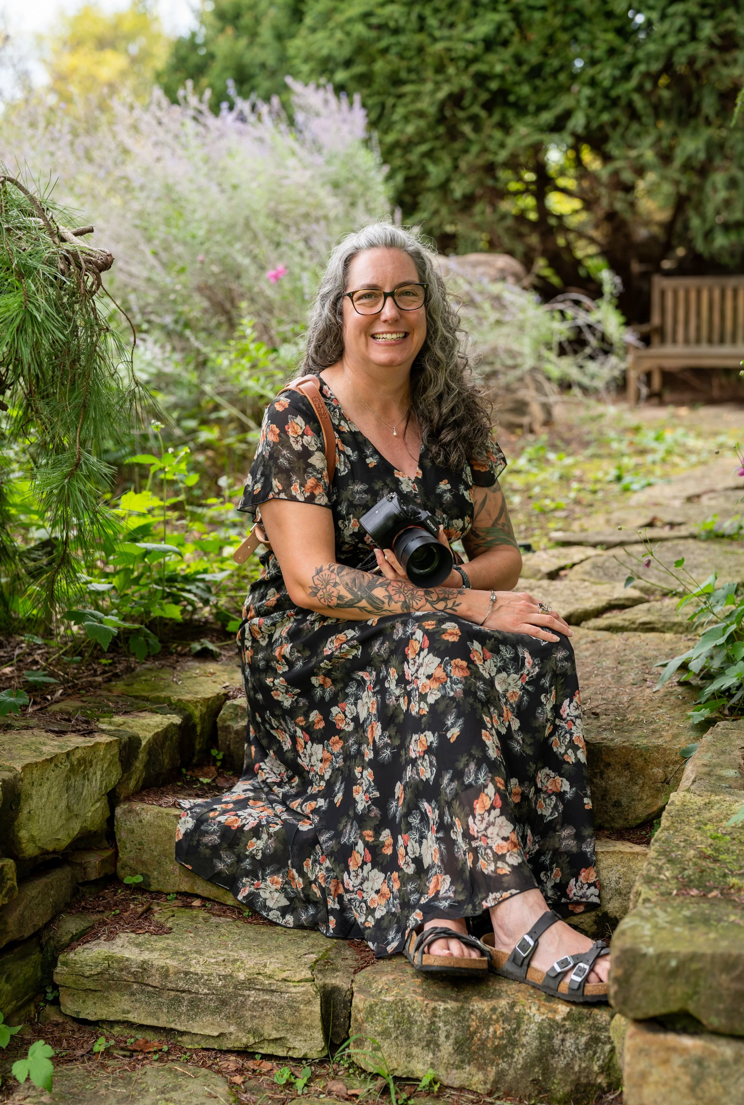 A smiling woman with curly gray hair, glasses, and tattoos on her arms, sitting on a stone ledge in a garden with green plants and trees in the background. She is holding a camera and wearing a floral dress and sandals.