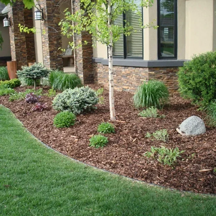 A landscaped garden bed with a variety of shrubs and plants, mulch, and a small tree in front of a house with large windows and brick accents.