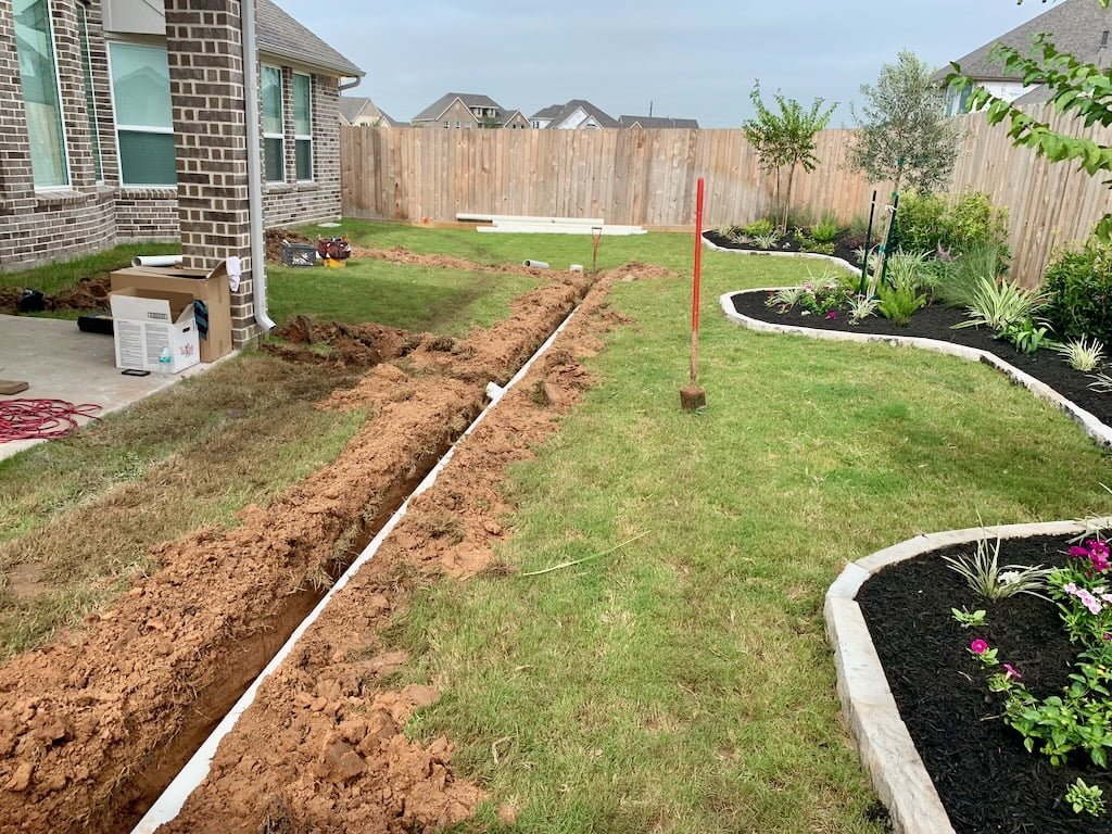 A backyard with a new flower bed bordering a lawn, with planting tools and a trench dug for installation, and a new wooden fence in the background.