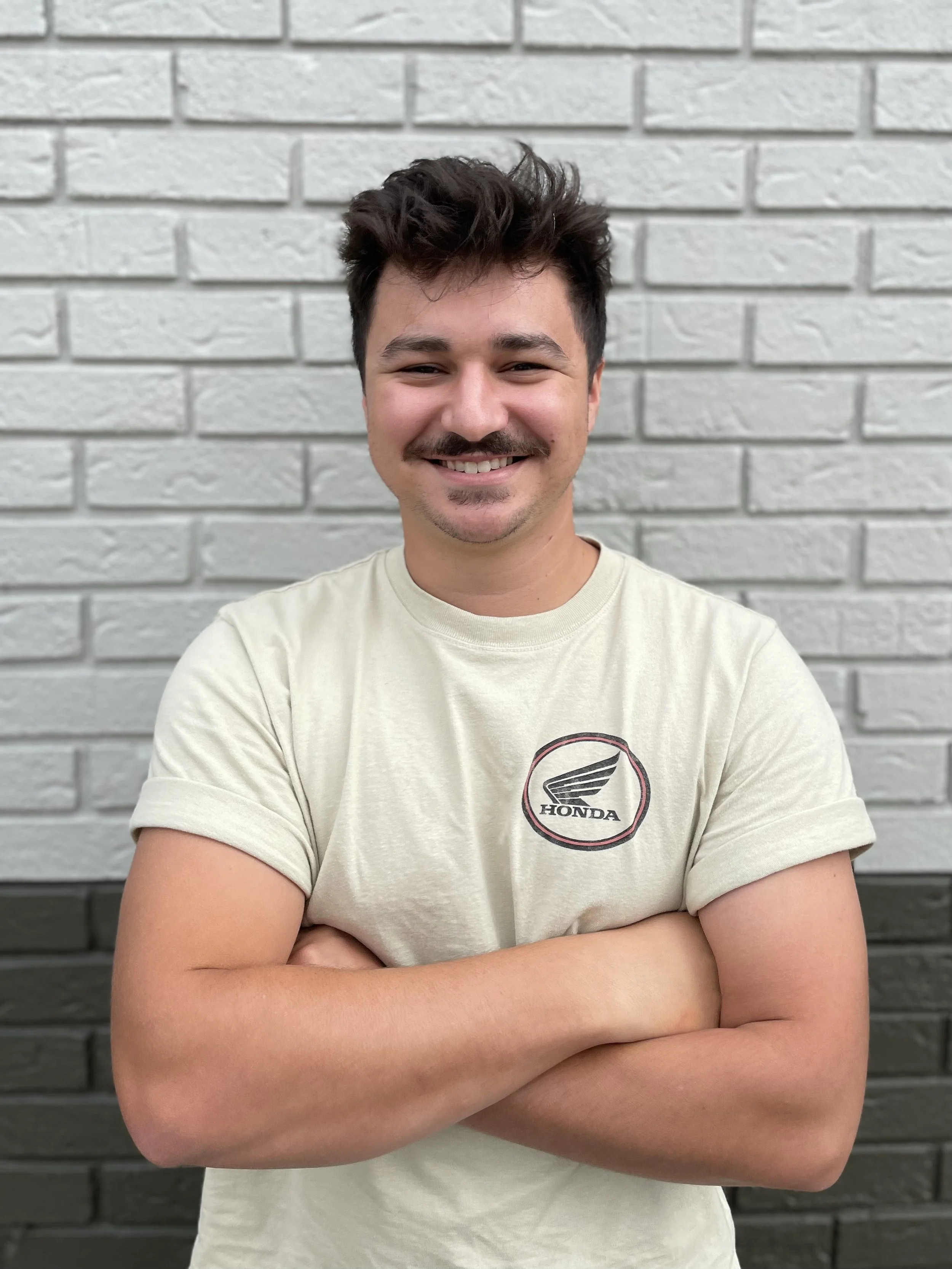 A young man with dark hair and a mustache smiling, standing in front of a light gray brick wall, wearing a cream-colored t-shirt with a Honda logo.