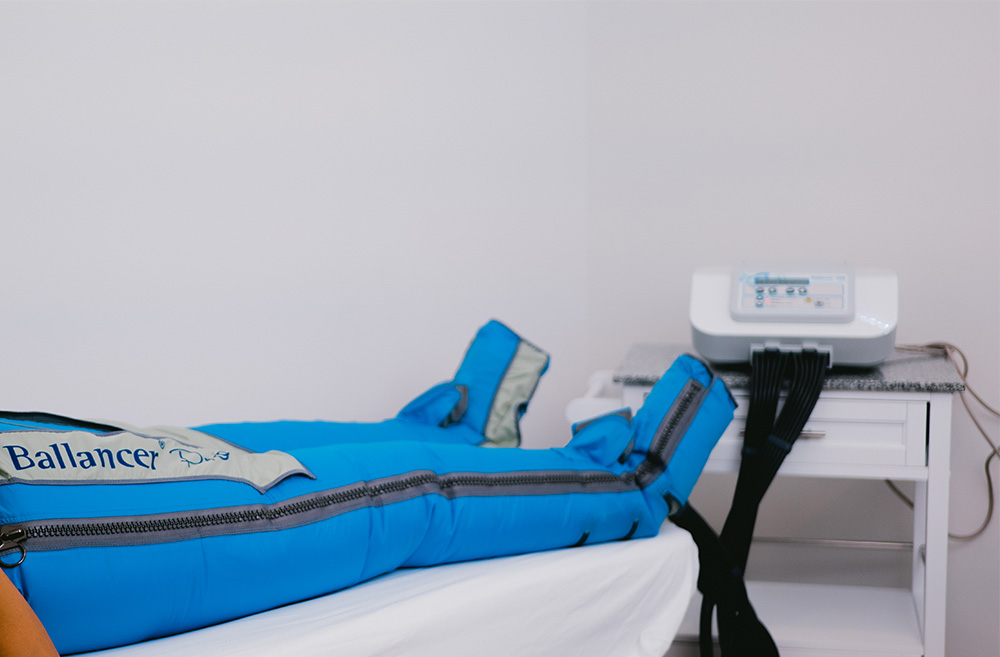 A person lying on a medical bed connected to a medical device, with the bed covered in blue padding and the device on a white table against a plain wall.