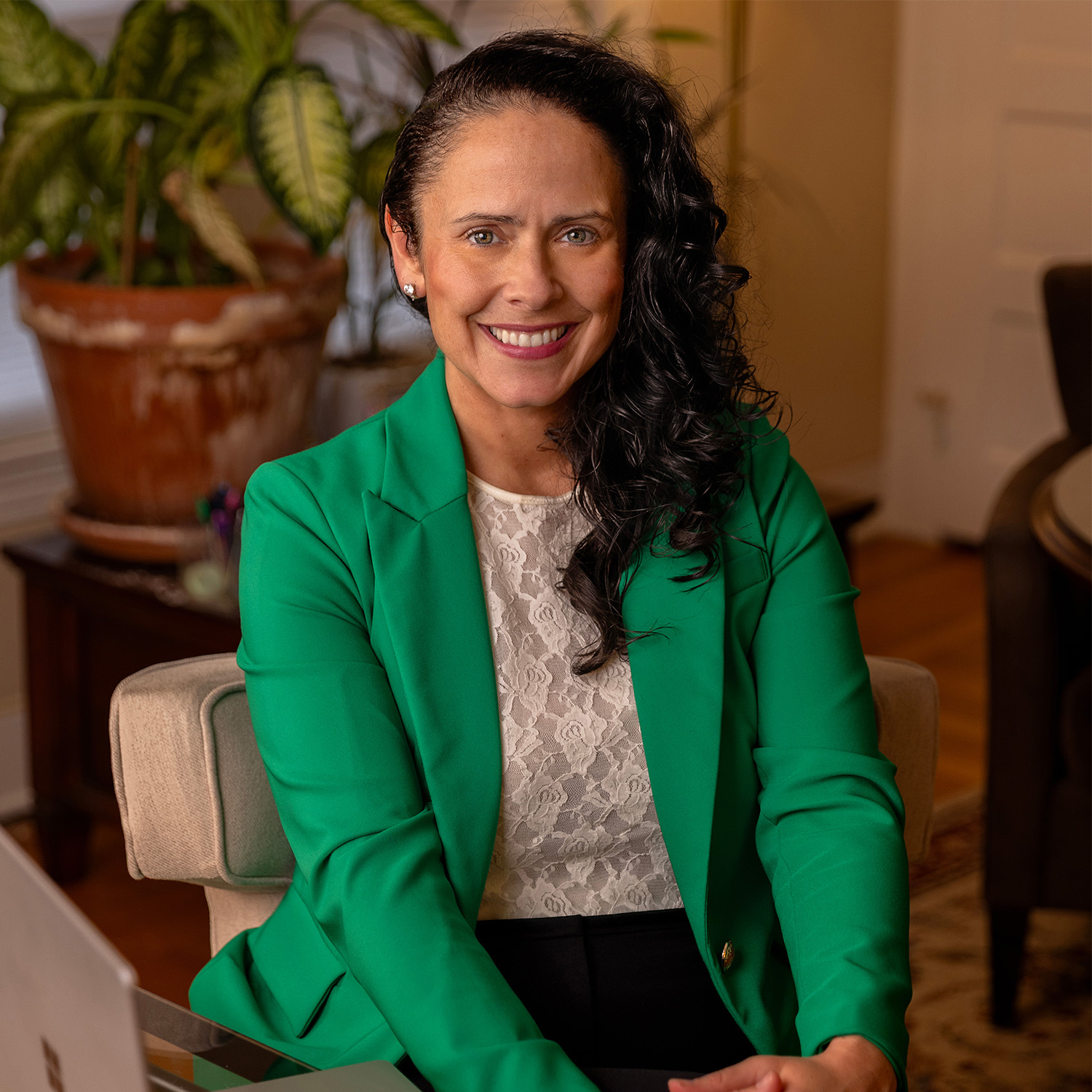A woman with curly dark hair wearing a green blazer and a white lace top, smiling and sitting at a desk in an indoor setting with a large potted plant in the background.