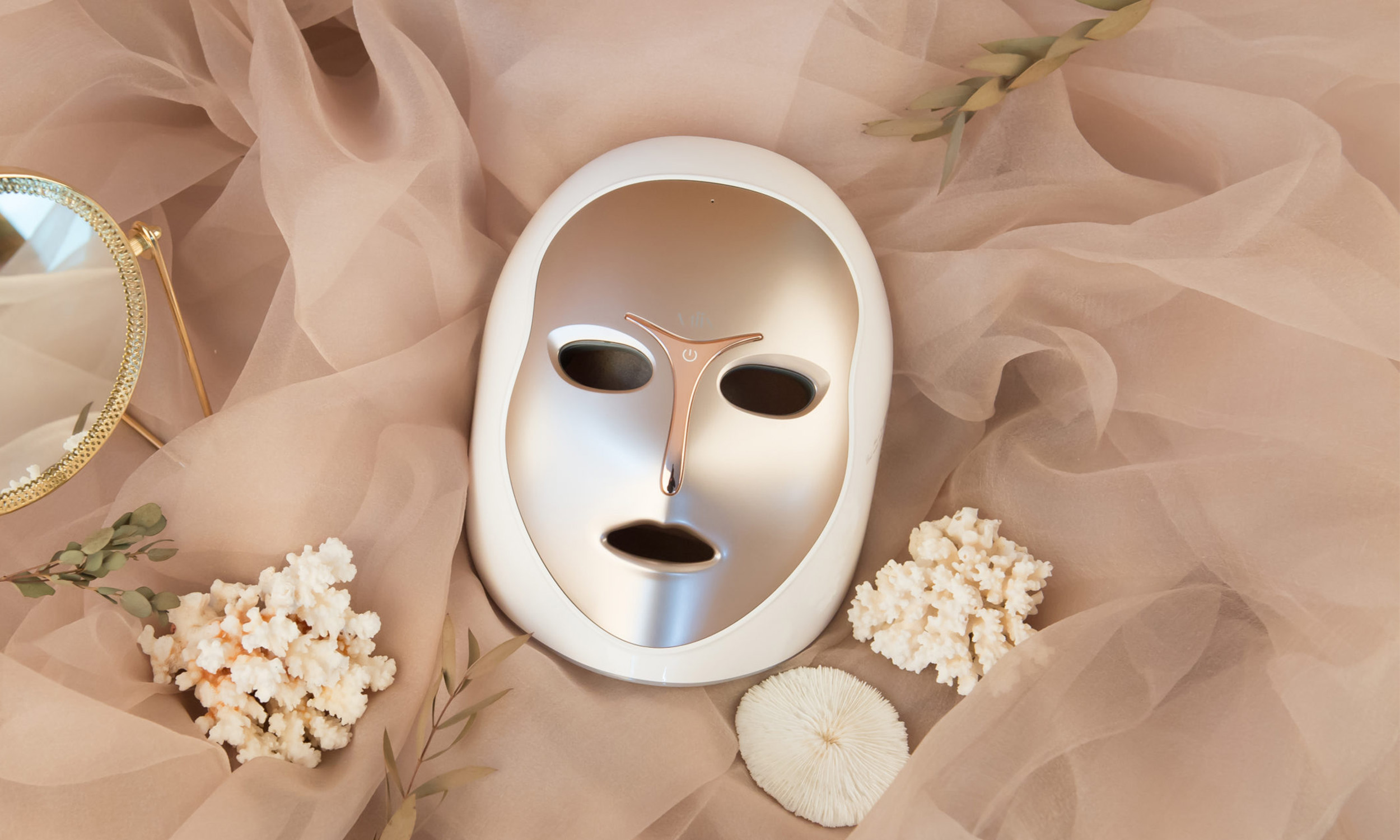 A white facial mask with cutouts for eyes and mouth lying on light pink, textured fabric surrounded by white dried flowers, a small round mirror with a gold rim, and green leaves.