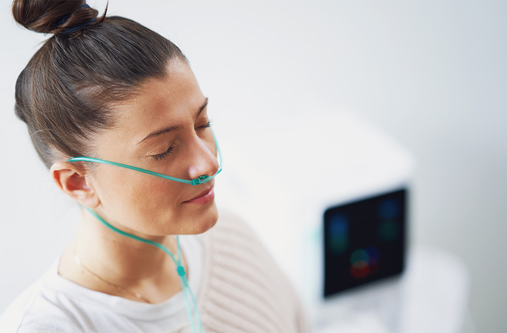 Young woman with a nasal oxygen tube resting on her nose and eyes closed in a hospital or medical setting.