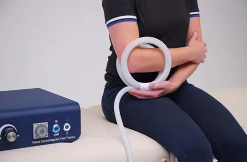 A person sitting on an examination table using a magnet therapy device on their arm, which is connected to a box with control knobs.
