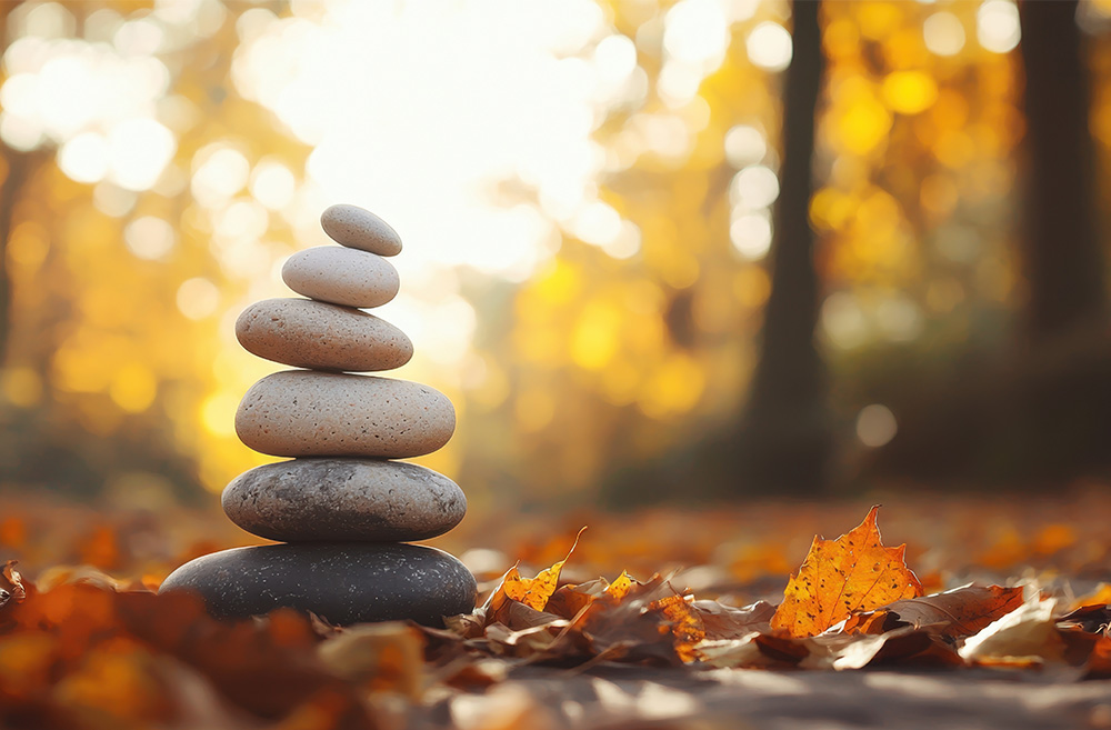 Stacked smooth stones arranged in a tower on the ground among fallen autumn leaves in a forest with blurred trees and warm light in the background.