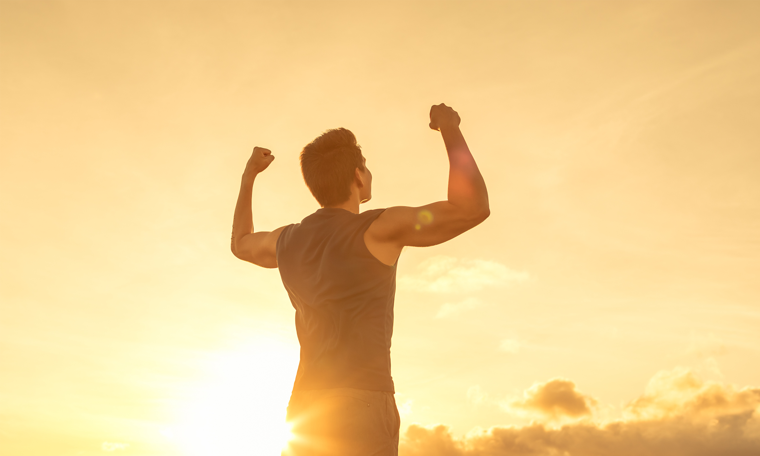 A male flexing his biceps in a tank top while the sun is setting.