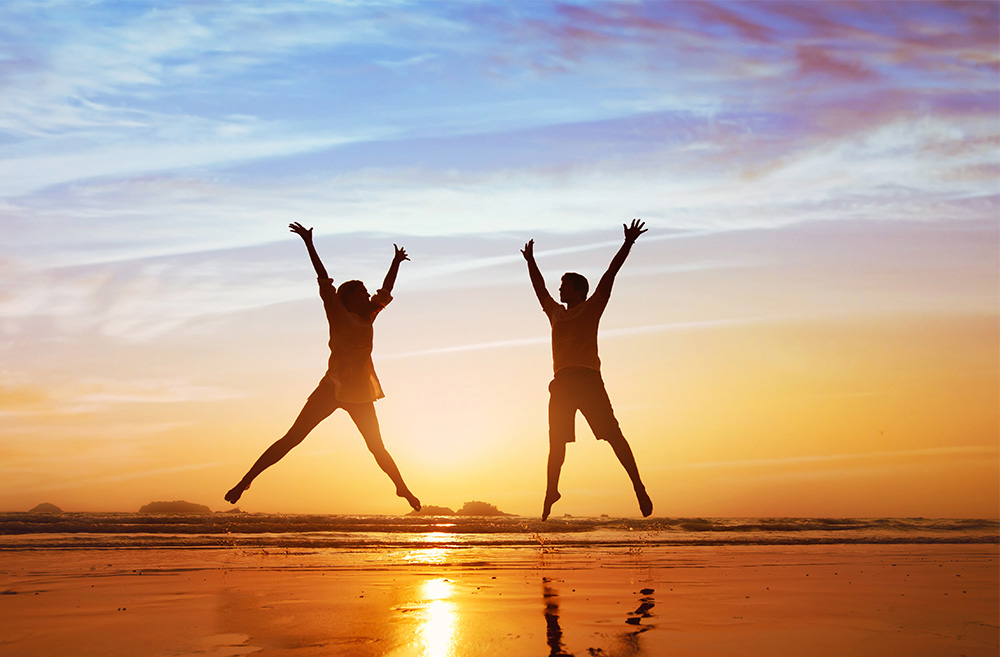 A male and female jumping at the beach while the sun sets over the ocean.