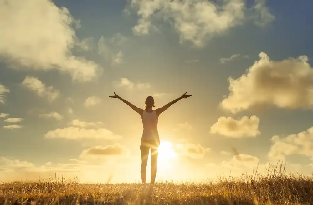 A person stands in a grassy field at sunset with arms outstretched, facing the sun with a partly cloudy sky in the background.