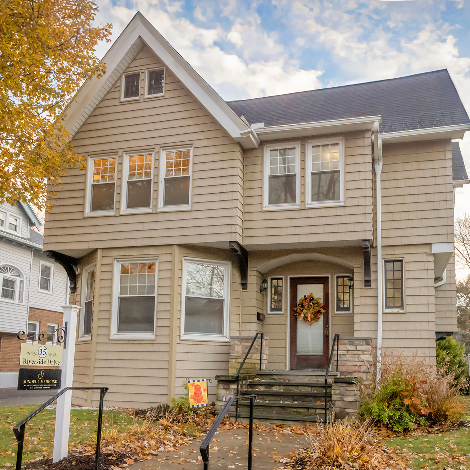 A Victorian-style house with beige siding, multiple white-framed windows, and a front porch with steps decorated with a fall wreath, surrounded by autumn foliage.