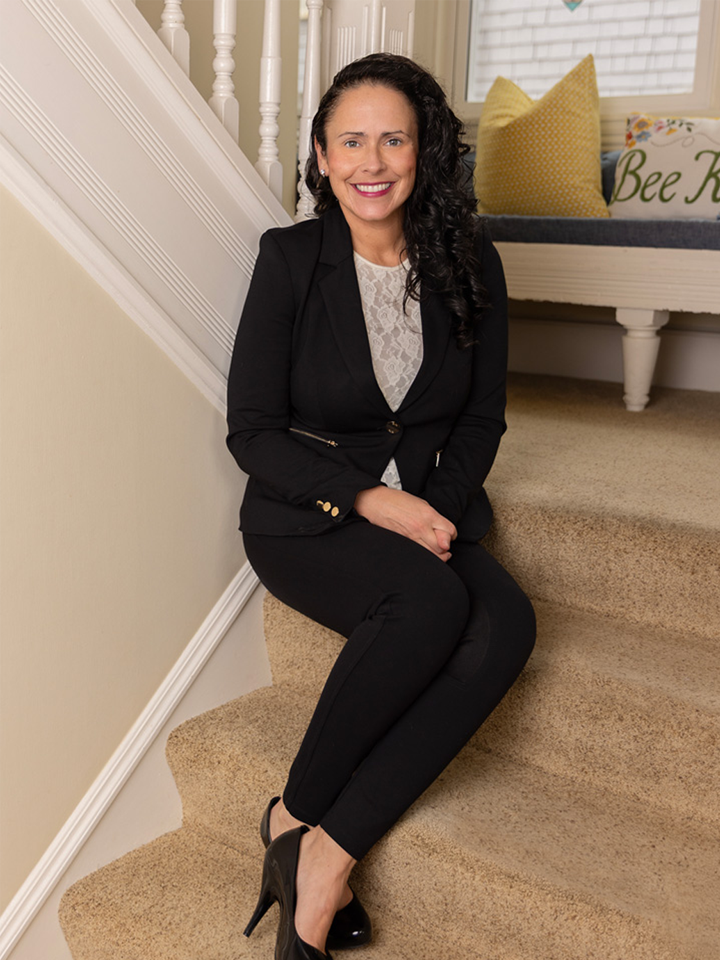 A woman sitting on carpeted stairs in a home, smiling, wearing a black blazer, lace top, black pants, and black high heels.