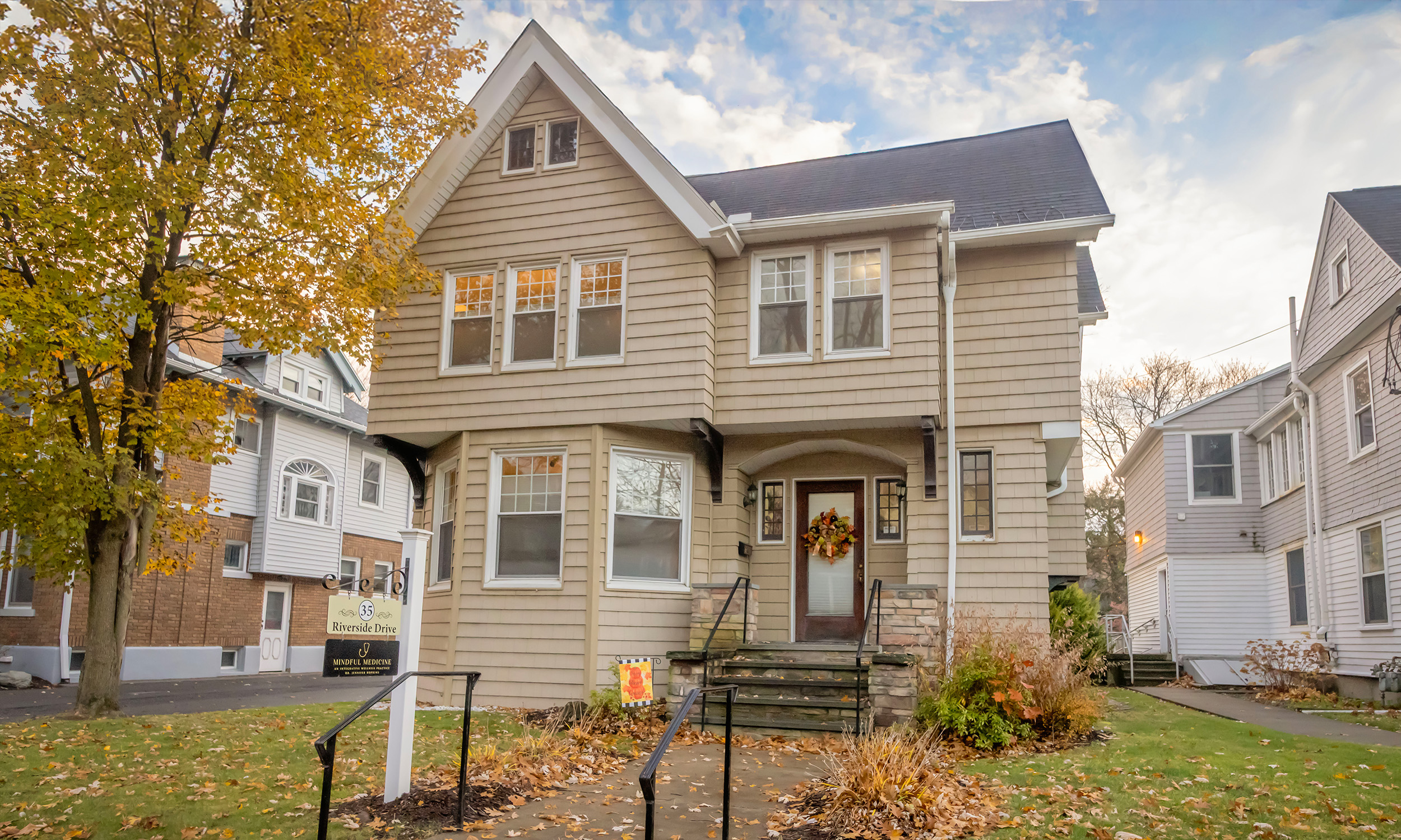 A beige, multi-story house with a front porch decorated with a fall wreath, surrounded by autumn leaves, with neighboring houses and a tree with yellow leaves on a cloudy day.