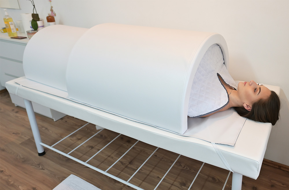 A woman lying on a bed inside a white oval-shaped tunnel machine, likely a phototherapy device, in a clinical setting.