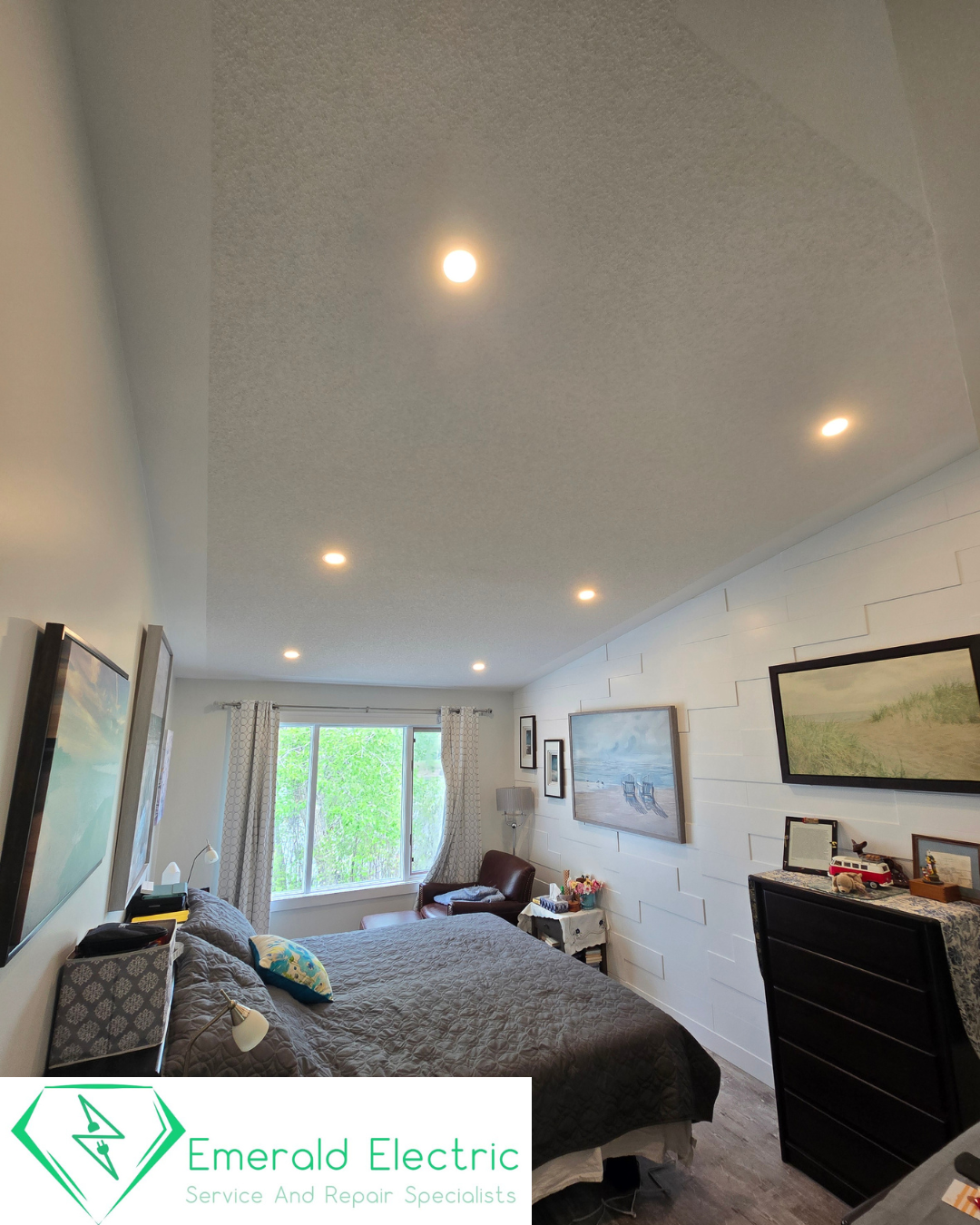 A cozy bedroom with a large bed covered in a dark quilt, a window with curtains, framed paintings on the wall, a small sitting area near the window, and various decorative items and furniture. The ceiling has multiple recessed lights.